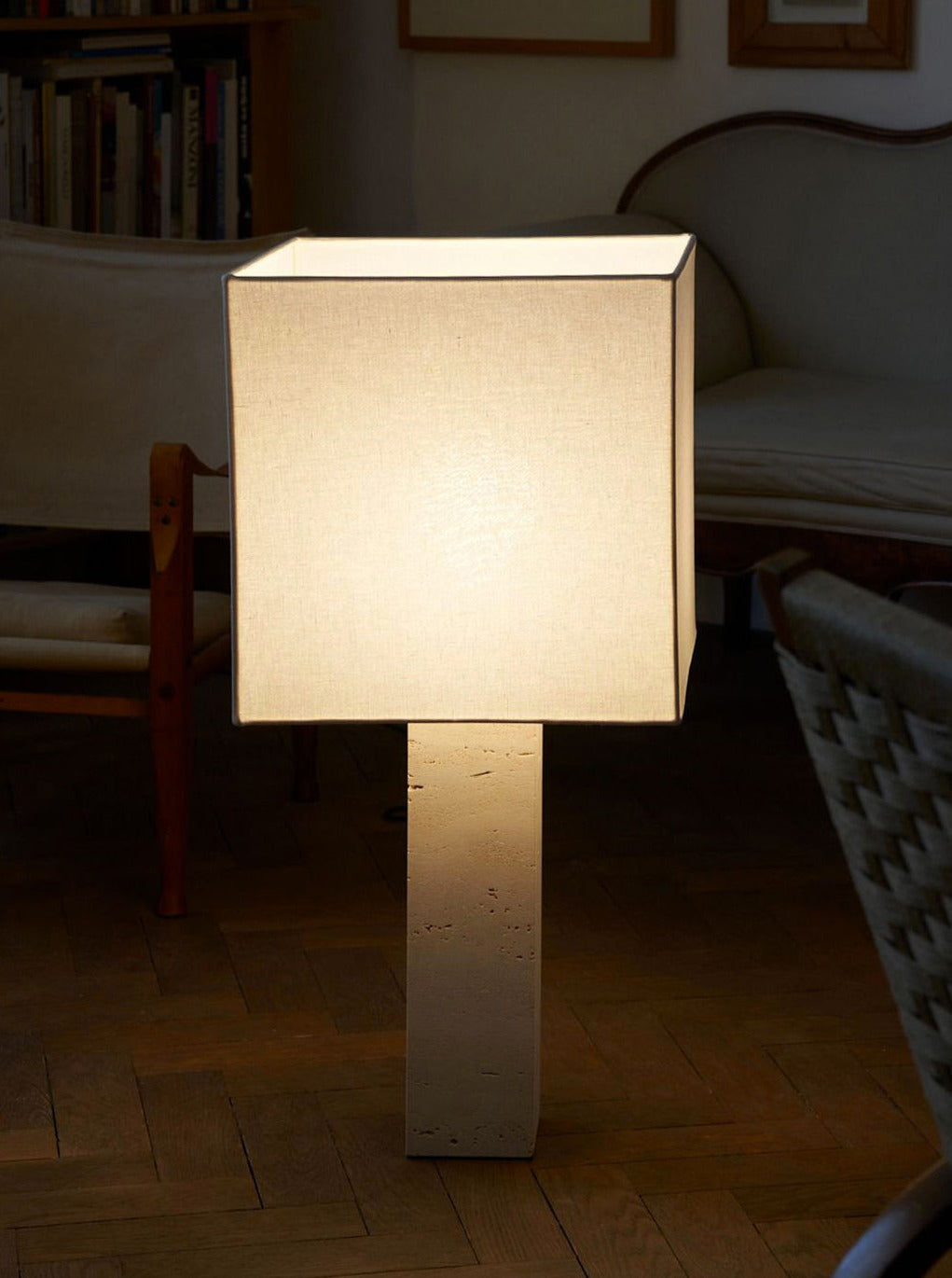 A modern, minimalist floor lamp with a beige square shade and a rectangular base glows softly in a cozy living room with wooden floors. The Nassi Lamps' Roma I Squared White Travertine casts a gentle light that complements the space beautifully. In the background, there is a beige armchair, a wooden bookshelf filled with books, and a beige couch, all embodying the essence of modernism.