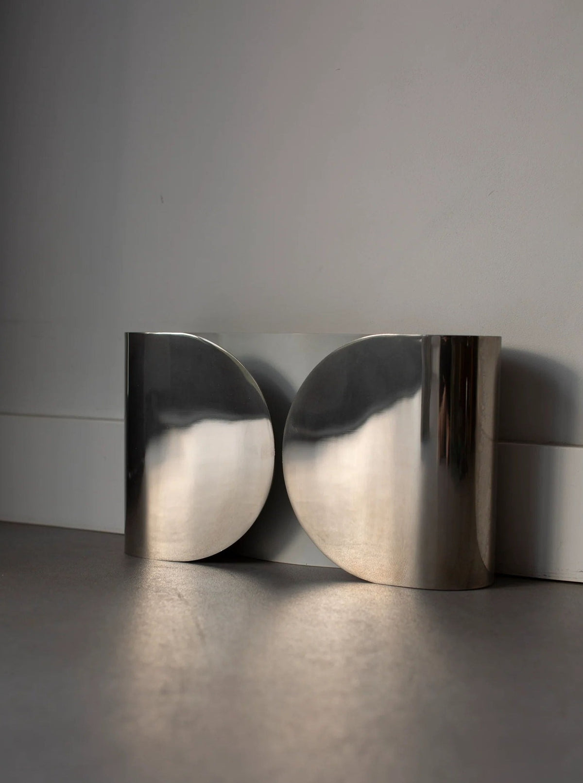 A sleek, modern sculpture sits against a plain backdrop. This polished metal piece, reminiscent of the Chrome Wall Lamp by Tobia Scarpa 80's from CU-RA-TED, features two large, convex circular shapes joined in the middle. It reflects light subtly off its smooth surfaces, casting gentle shadows on the surrounding surface.
