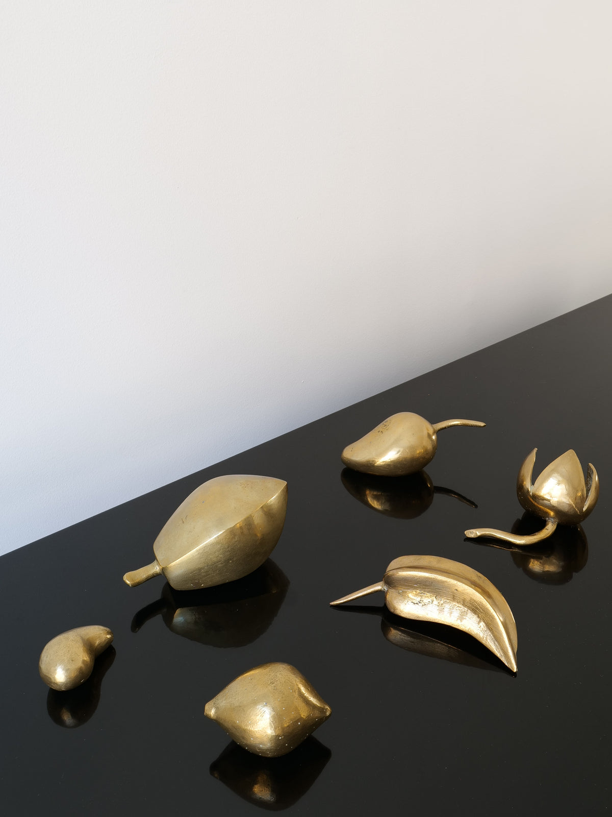 A selection of small, solid brass sculptures, part of the Brass Sculpture Collection by Collection apart, is gracefully arranged on a glossy black surface. The backdrop showcases a simple, light-toned wall that adds a touch of midcentury French artistry.