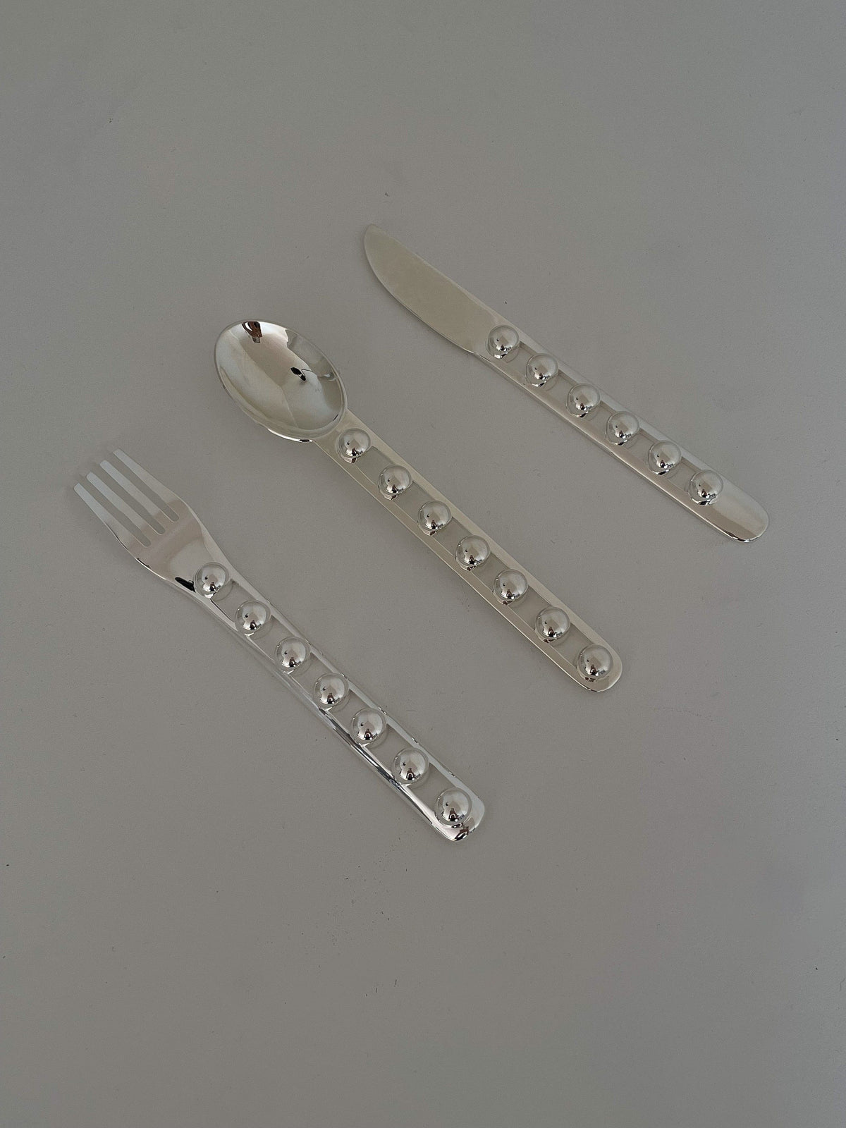 The "Seven Ball" Cutlery Set by Jessi Burch is displayed on a gray background, highlighting its functional art that includes a fork, spoon, and knife. Each piece boasts uniquely textured handles decorated with raised circular designs.