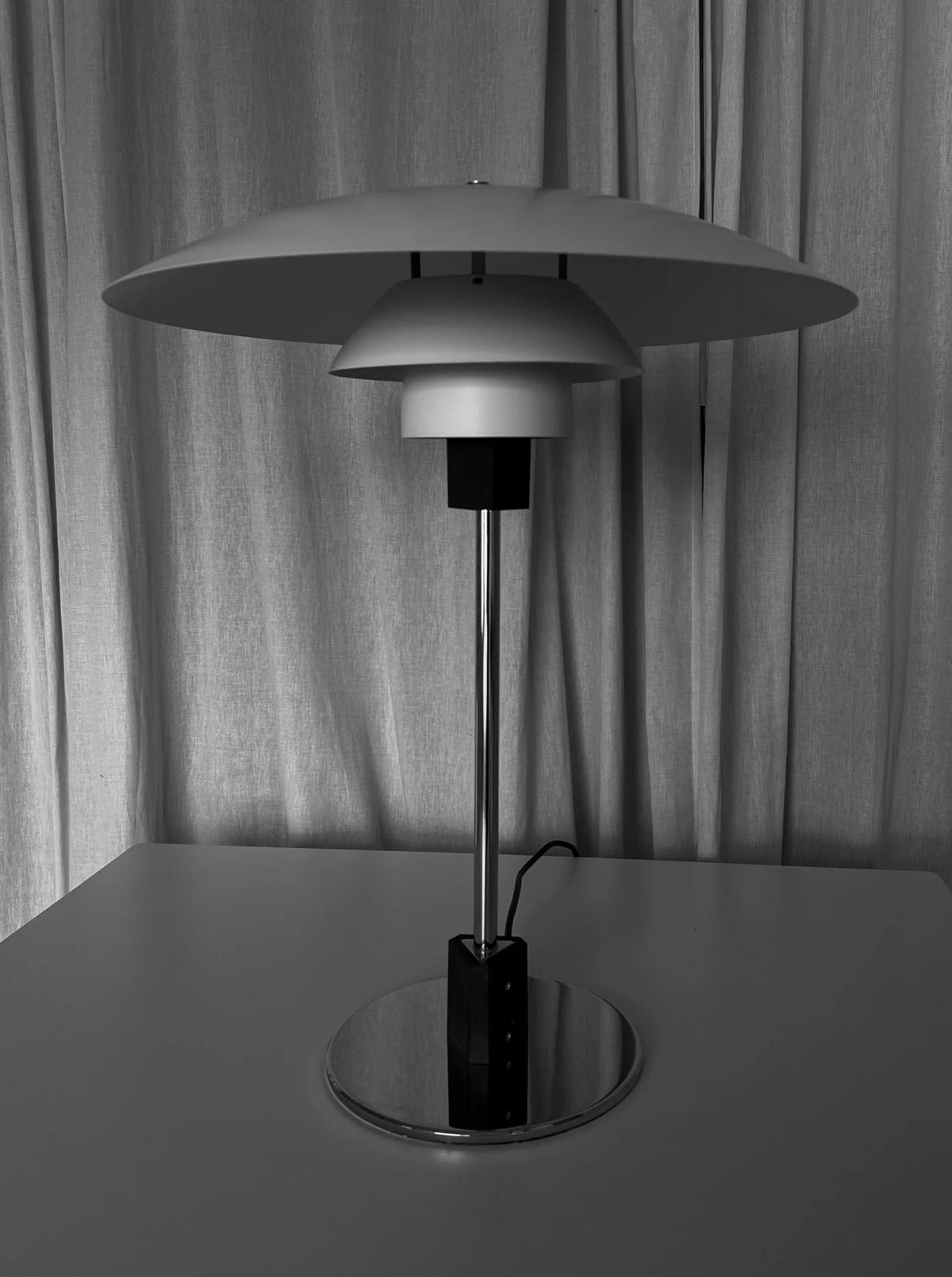 A black and white image of a modern, vintage table lamp on a flat surface. Resembling Poul Henningsen's iconic designs, the CU-RA-TED Poulsen Table Lamp PH 4/3 1983 features a wide, disc-shaped shade and a slim, tall stem mounted on a circular base. A cord extends from the base while curtains hang in the background.