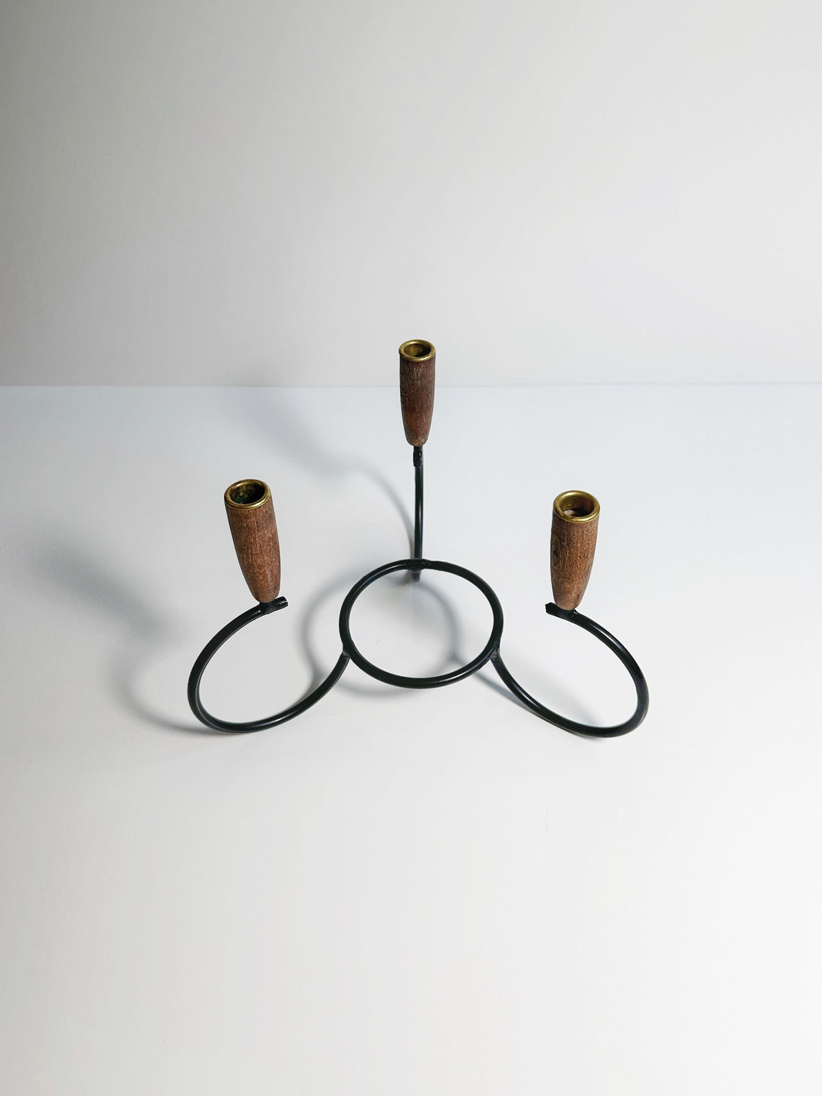 The Danish Candleholder 1960s from Dodo Vintage features a minimalist black metal design with three circular bases and wooden candle cups finished with teak ends. The sleek and simple structure is elegantly displayed against a plain, light grey background.
