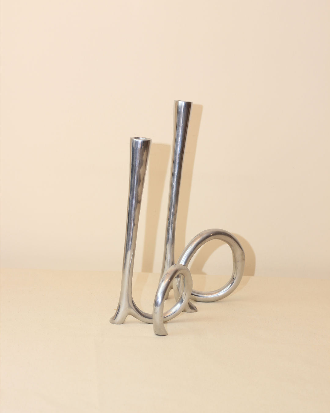 The Cast Aluminum Candleholders by Boga Avante Shop feature modern silver looped bases and tall stems, perfect for minimalist decor, displayed on a beige surface with a neutral backdrop.