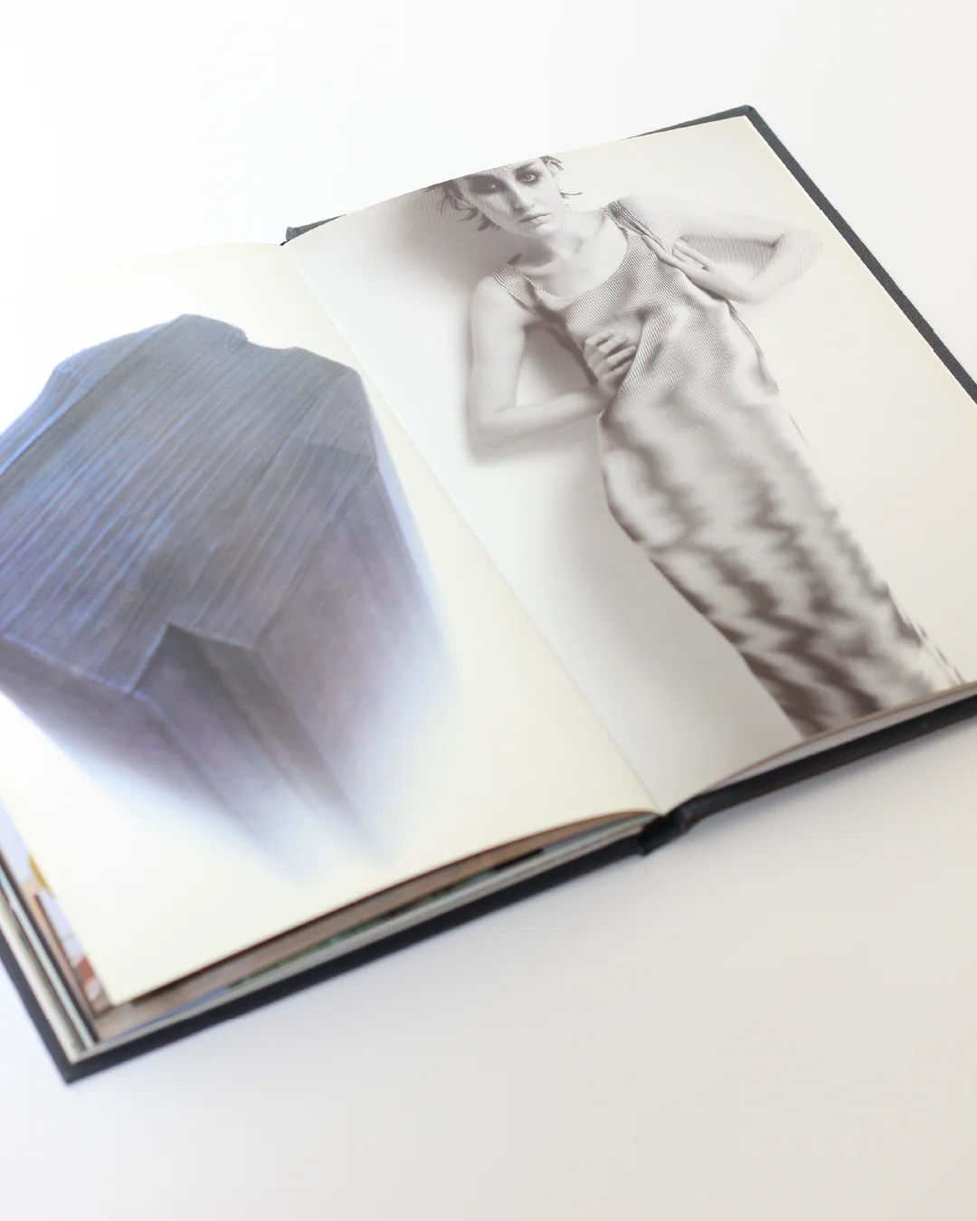 The Issey Miyake Book by Boga Avante Shop is open on a white surface, displaying a blurred grayscale photograph of a person in a patterned dress on the right page, evoking Issey Miyake's iconic pleating techniques. The left page presents an abstract geometric shape that mirrors contemporary fashion trends.