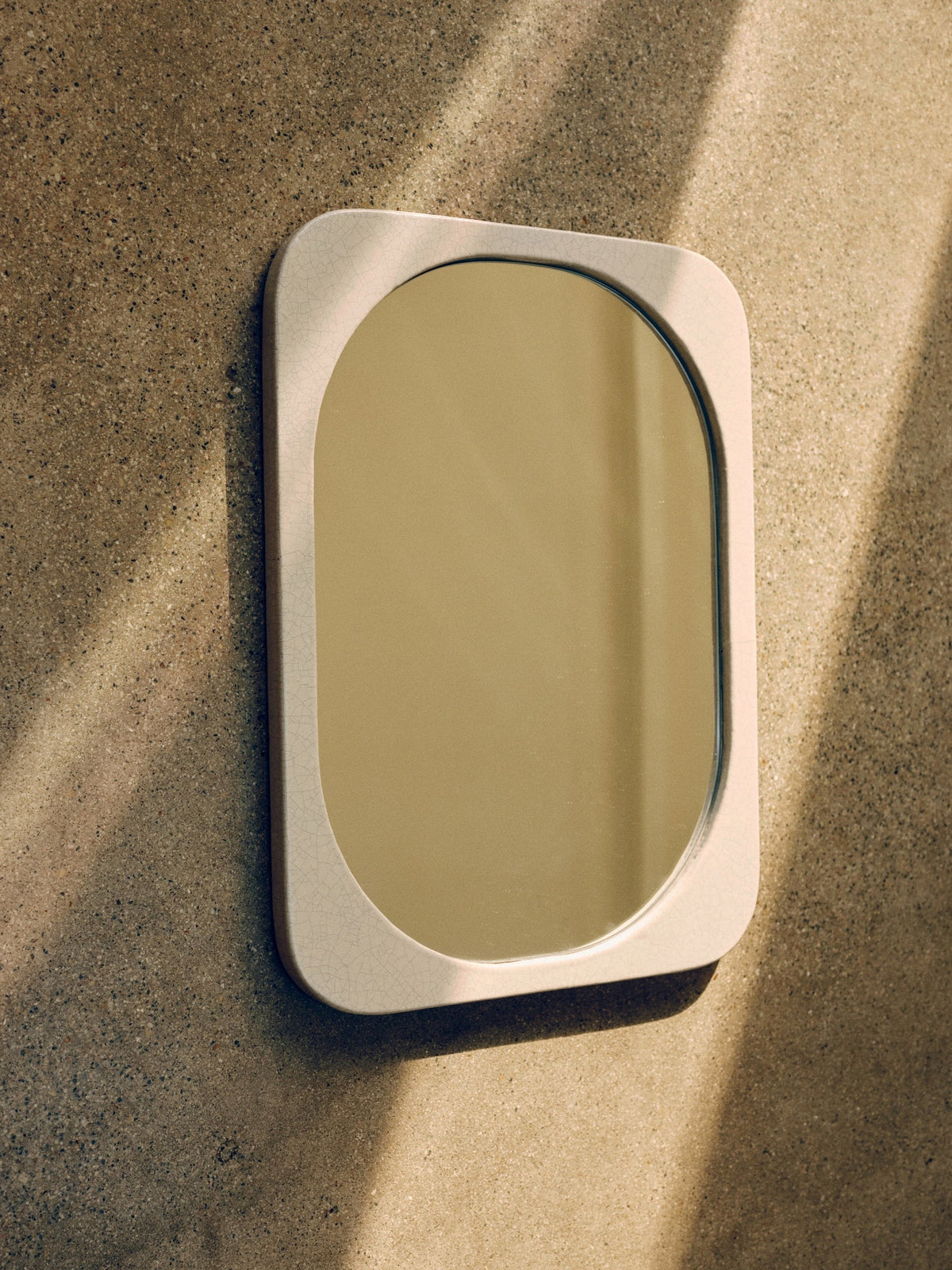 A wall-mounted Mirror Bien Fait in Lava Stone by Bien Paris with a light-colored rectangular frame and a crackled enamel finish is set against a textured, beige wall. Sunlight streams in, casting diagonal shadows across the mirror and wall. Made in Italy, this elegant piece enhances any space with its refined craftsmanship.