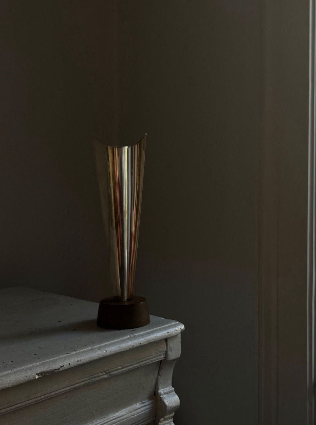 A sleek Médecine Tapio Wirkkala Silver Vase with a narrow neck stands on a distressed, vintage white dresser. The background is dark, creating a contrast that highlights the simplicity and elegance of the vase against the aged texture of the dresser.