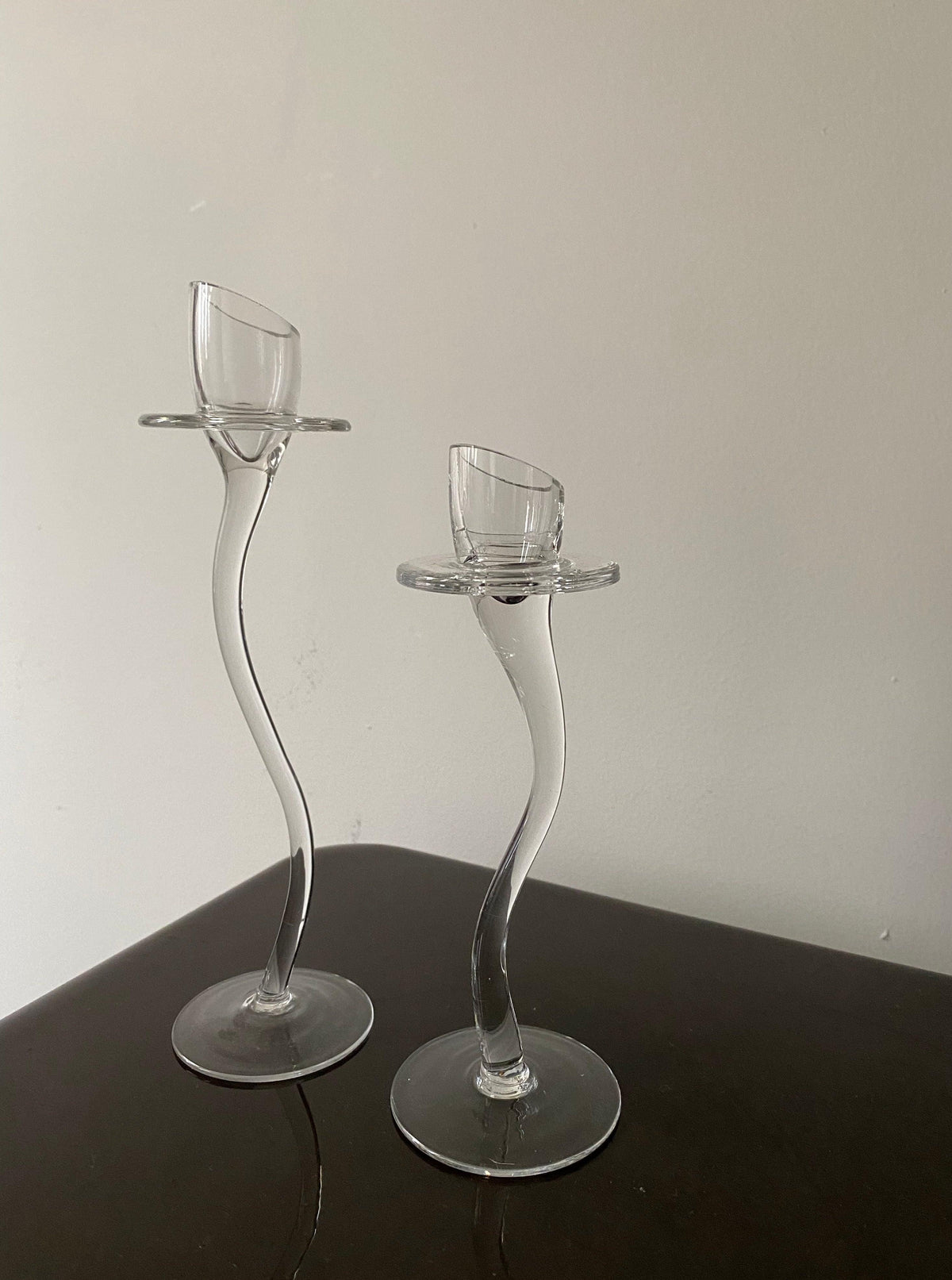 Two elegantly curved Les Objets Amsterdam clear glass candle holders with votive cups, exuding refined elegance, standing on a dark surface against a light gray background.