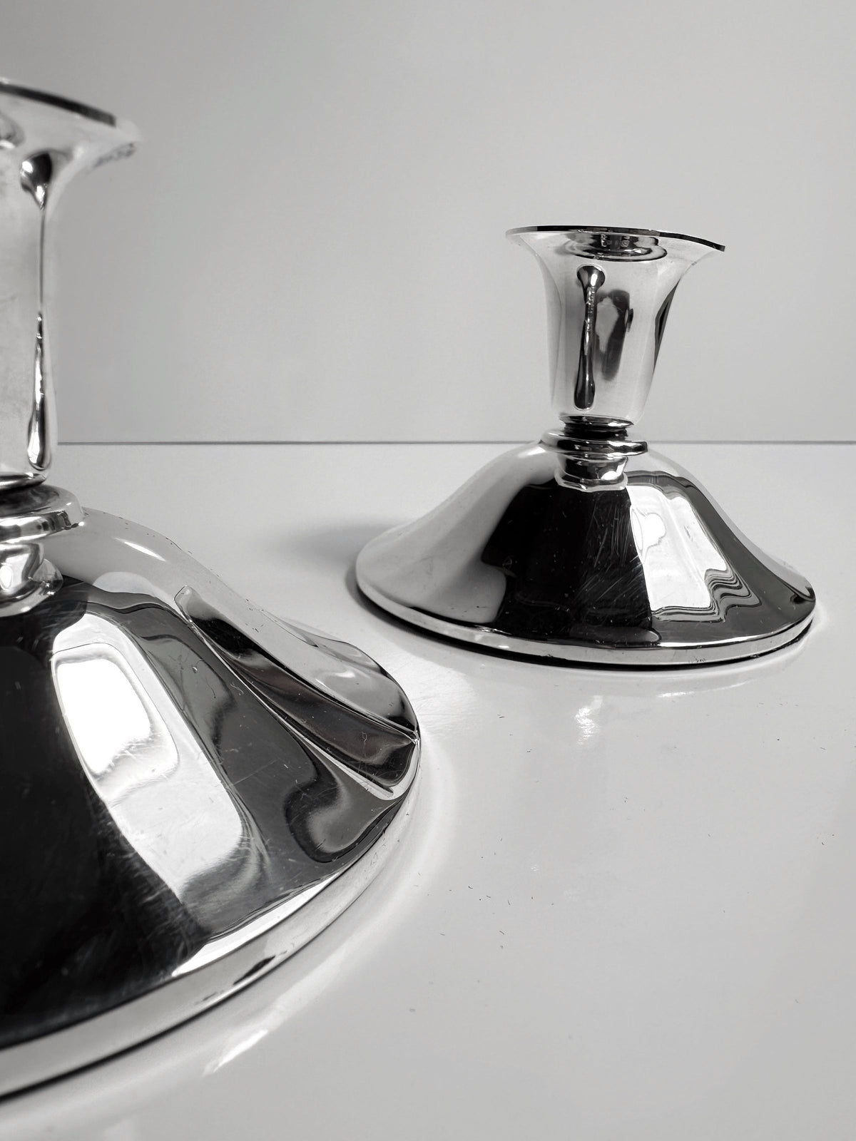 Close-up of the Silver-Plated Art Deco Candleholders - 1930 by Dodo Vintage, positioned on a glossy white surface against a plain background. These candleholders feature a sleek, modern design with wide bases and narrow tops. Their reflections enhance an elegant, minimalist aesthetic that evokes classic Art Deco style.