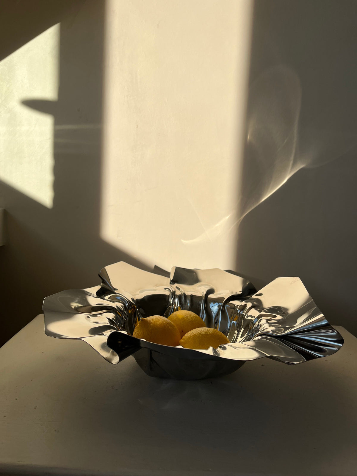 A Folded Chrome Bowl from septembre studios, with its angular design, holds three lemons and is set on a white surface. Soft sunlight and shadows create a warm ambiance in the room, casting shapes on the wall behind it.