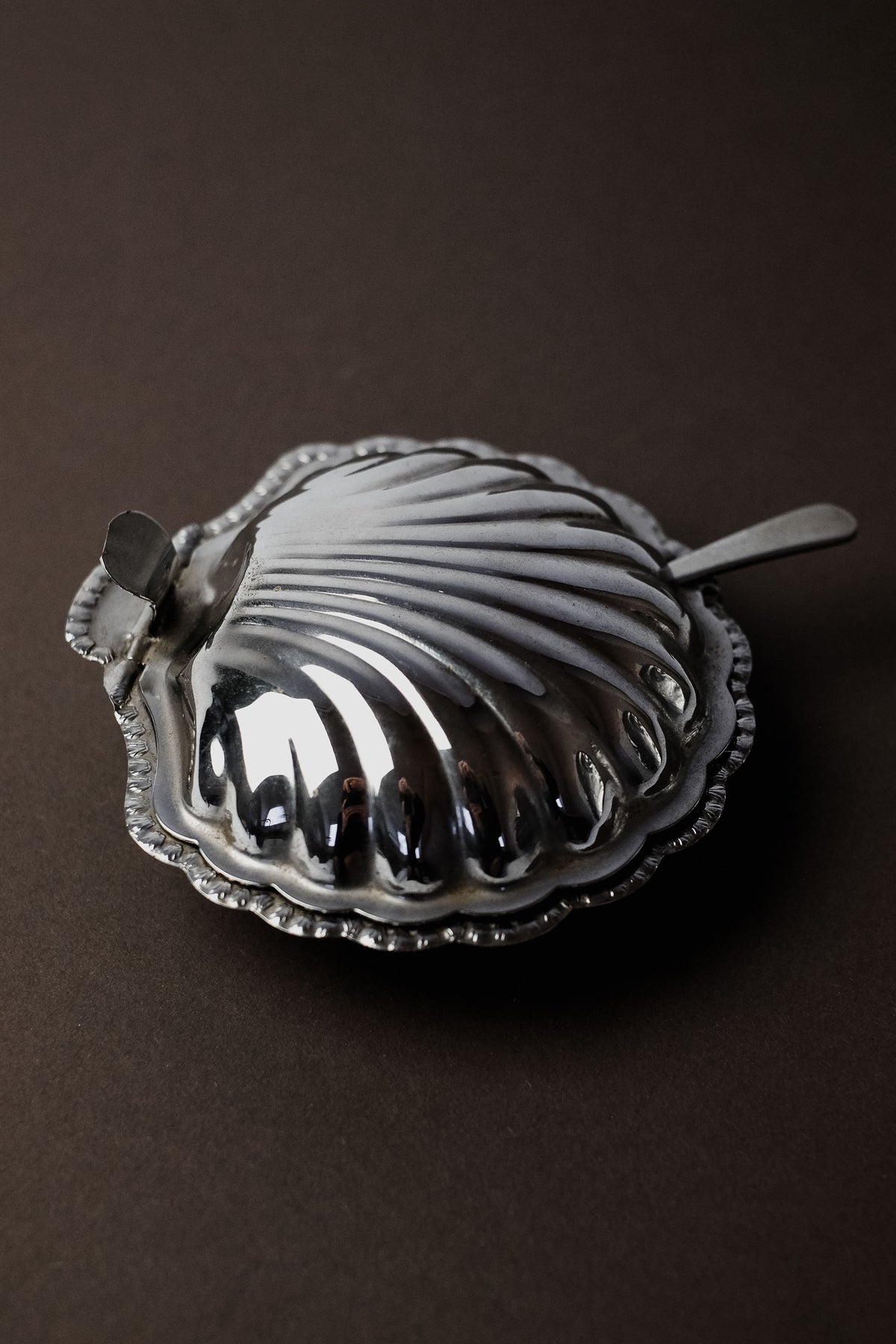 The Shell Shaped Butter Dish with Spoon by septembre studios is a shiny, silver seashell design featuring intricate ridges and scalloped edges. Perfect for an exquisite dining experience, it's completed with a matching lid and rests elegantly on a dark surface.