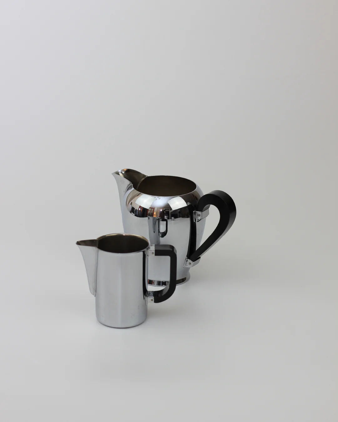 Two shiny metal pitchers with black handles from the Boga Avante Shop’s Italian Coffee and Tea Set, 1945, sit on a plain light gray surface; the larger pitcher is positioned behind and to the right of the smaller one.