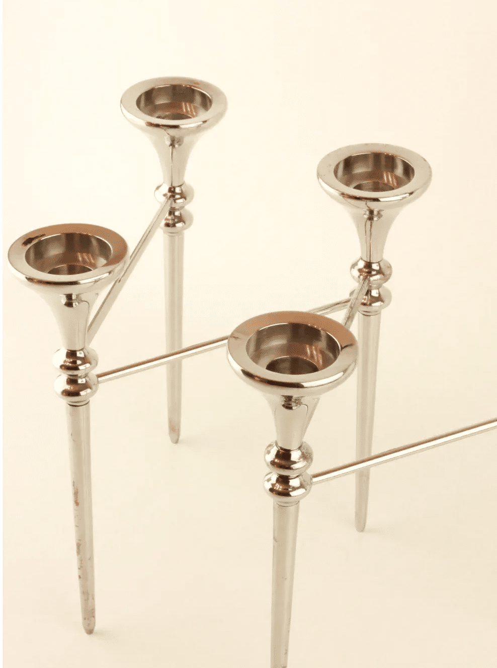 A sleek, four-branched, modern silver candlestick holder with a minimalist design. The Scandinavian Modular Candelabra from Boga Avante Shop features four elevated cylindrical candle cups atop slender, conical stems connected by modular arms. The polished metal finish evokes a subtle nod to a vintage chrome chandelier.