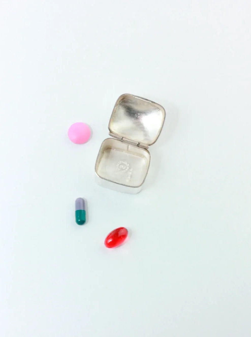 A beautiful handcrafted Silver Pillbox With Onyx Stone from Boga Avante Shop, created in Taxco, Mexico, is displayed open alongside three varieties of pills: a pink round one, a two-toned green and purple capsule, and a red oval pill—all set against a plain white background.
