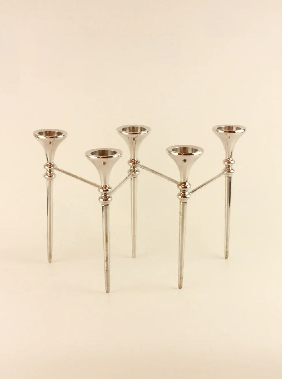 The Scandinavian Modular Candelabra by Boga Avante Shop is a modern, metallic candelabra with five candle holders arranged in a symmetrical design. The holders are elevated on slender, modular arms connected to a central frame, which has three legs for support. The overall aesthetic is sleek and contemporary, reminiscent of a vintage chrome chandelier.
