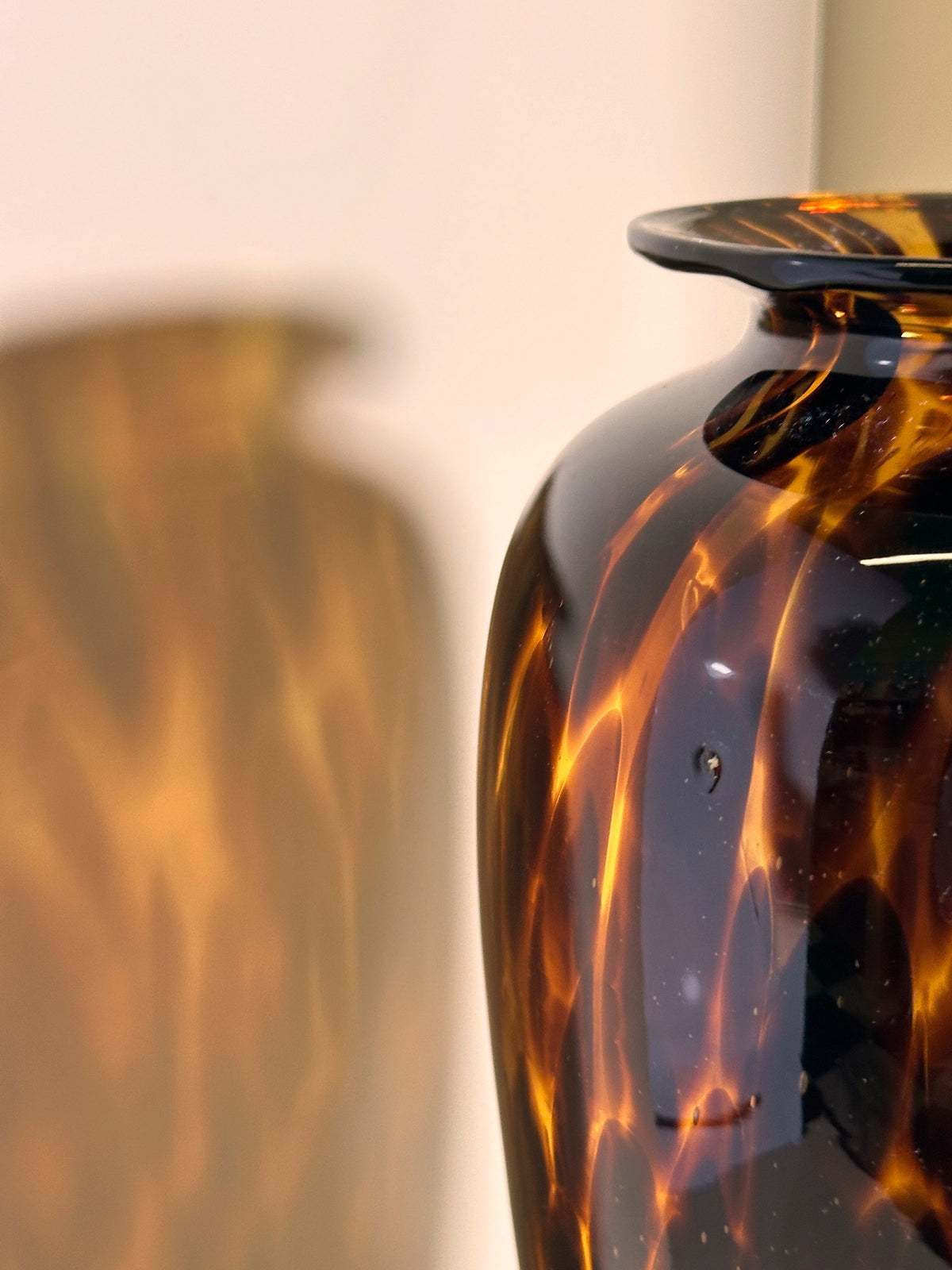 Close-up of the glossy Tall Murano Tortoise Vase by Dodo Vintage, featuring an amber marbled pattern that reflects light smoothly. The left-side reflection creates a soft echo of its design, reminiscent of Tortoise speckle technique, with a softly blurred background.