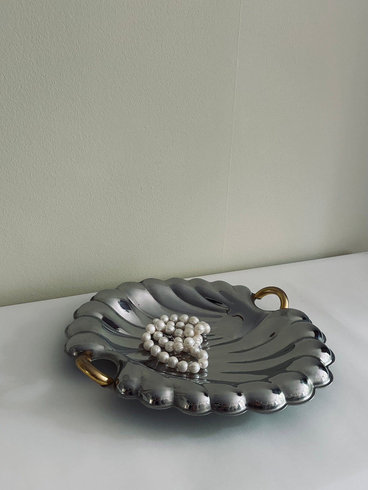 Crafted in Italy, the Flock Studio Italian Stainless Steel Shell Tray features a luxurious design with scalloped edges and two gold handles. It elegantly showcases a string of pearls arranged in a heart shape, resting on a white surface against a plain beige wall background.