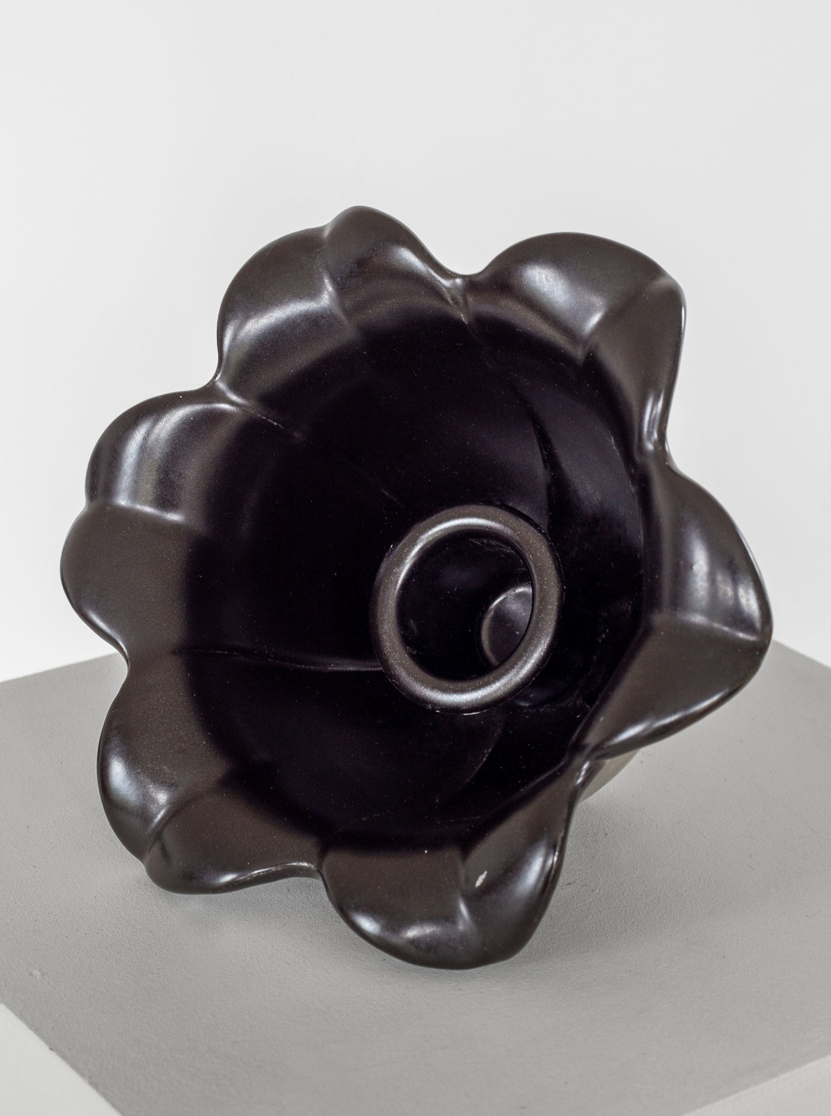 A dark, scallop-edged ceramic bowl with a glossy finish rests on a light gray pedestal. Reminiscent of Spigel's Swedish Mid-Century Modern studio pottery from the 1950s, the bowl features a circular raised ring in the center, adding an element of dimension to its smooth and symmetrical design.