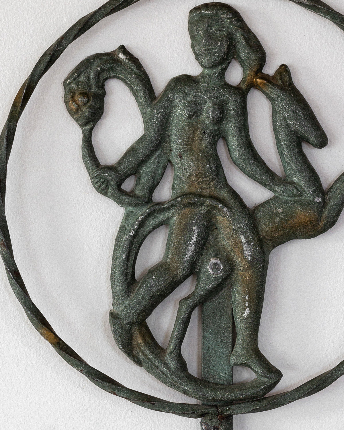 A close-up of an ancient-looking wrought iron sculpture by Spigel shows a human figure entwined with two horse-like creatures, encircled by a thin metal ring. It evokes the Art Deco style of the 1920s/30s Swedish wall sconce, set against a plain white background.