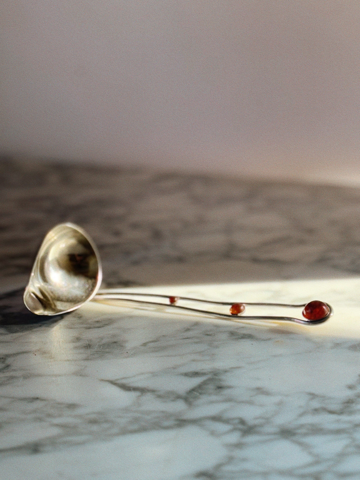 The Huerto Silver Plated & Amber Insert Serving Ladle, featuring a curved handle and two amber accents, crafted by a Polish artisan, rests on marble with soft light casting gentle shadows.