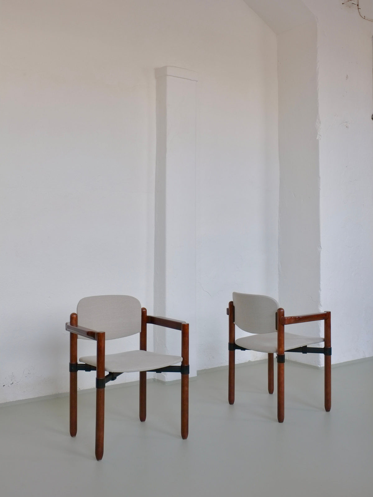 Two Fröscher Sitform stacking armchairs by Veter Vintage, featuring wooden frames and light gray cushions, rest side by side against a white wall on a smooth floor. Their minimalist elegance and clean lines echo the 1970s design from the set of four armchairs.