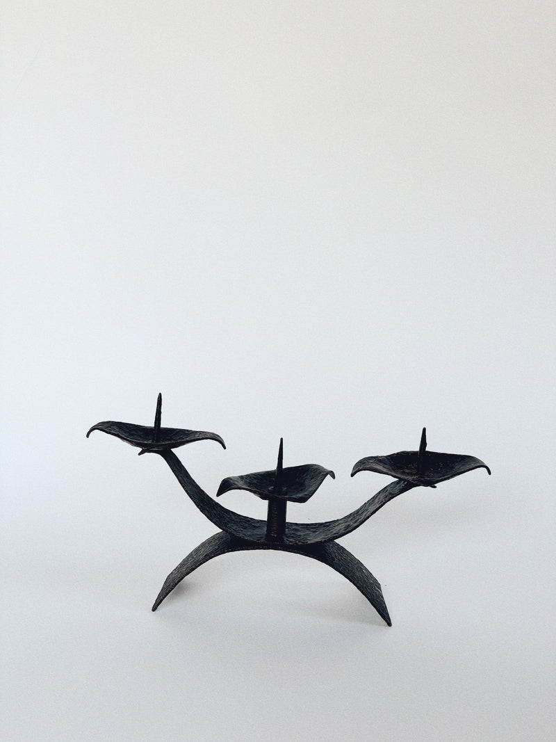 The Huerto 3 Arm Metal Candleholder, designed with sleek, curved lines in minimalist black metalwork, features three prongs for individual candles against a plain white background. It's artisan-made with a modern style.