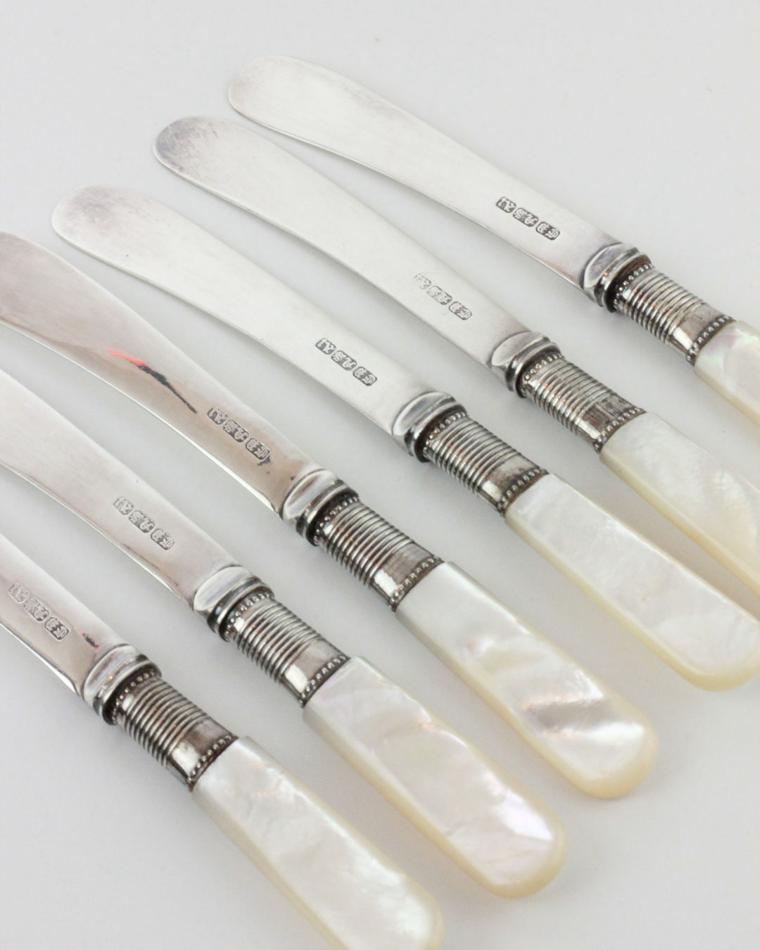 Set of six Boga Avante Shop Mother-of-pearl Butter Knives, each with English silver blades, hallmarks, ornate bands, and pearlescent handles, elegantly arranged diagonally on a white surface.