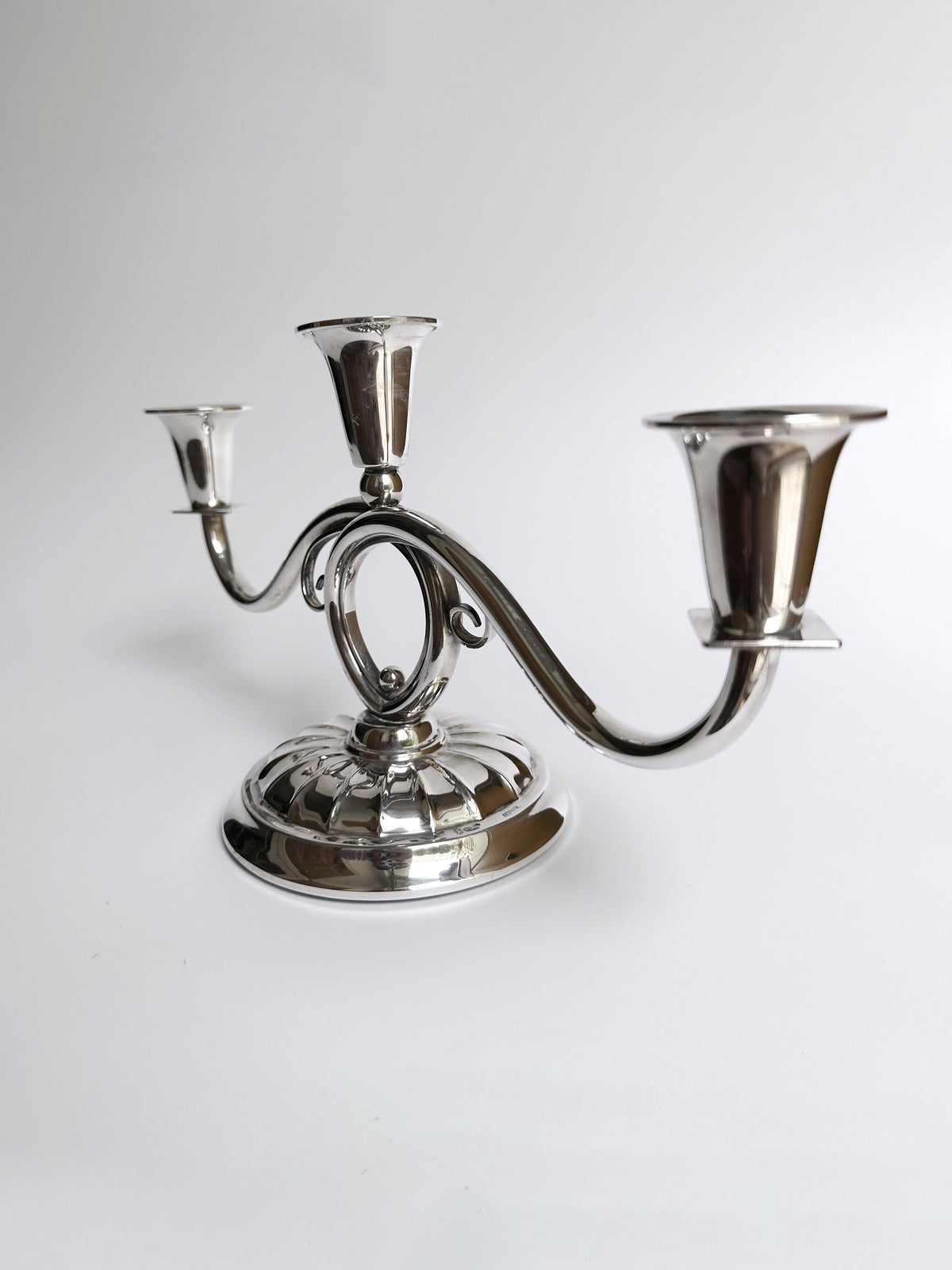 The Danish Mid Century Candleholder from Dodo Vintage, featuring a silver-plated finish and an ornate filigree design, is set against a plain white background. The central arm is slightly elevated above the other two, each of which holds a candle holder. The base is round and reflective.