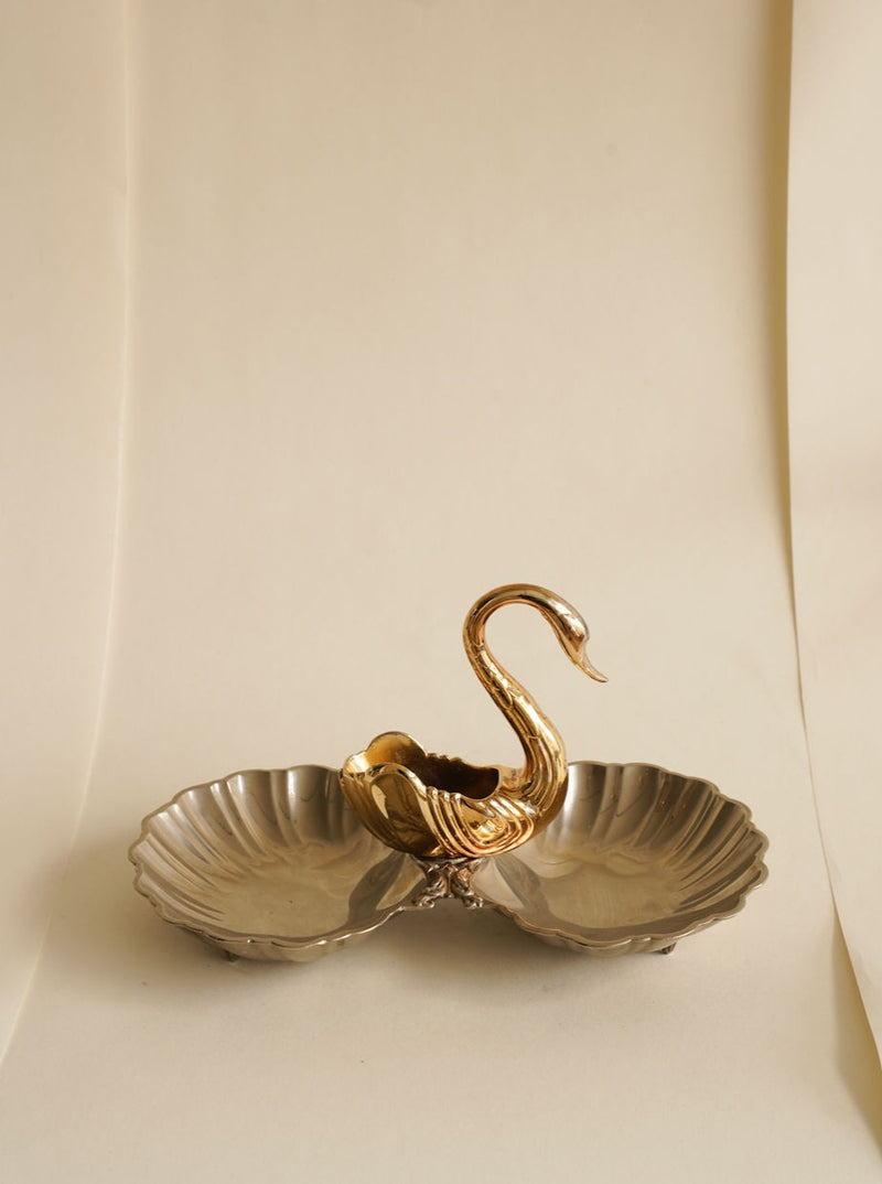 A vintage serving dish featuring a dual-compartment design shaped like clam shells, with a central handle in the form of a golden swan. This elegant piece stands on a neutral background, highlighting its intricate and timeless swan decoration. It's the Shell-Shaped Swan Serving Dish by Les Objets Amsterdam.