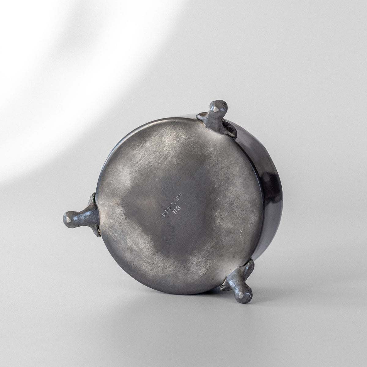 The vintage Spigel Art Deco Metal Box from 1934, with its round shape and three short legs, rests upside down on a softly lit white surface. Its underside features a weathered appearance with some imperfections and a faint central inscription, evoking the essence of an antique pewter pot. The background remains plain and understated.
