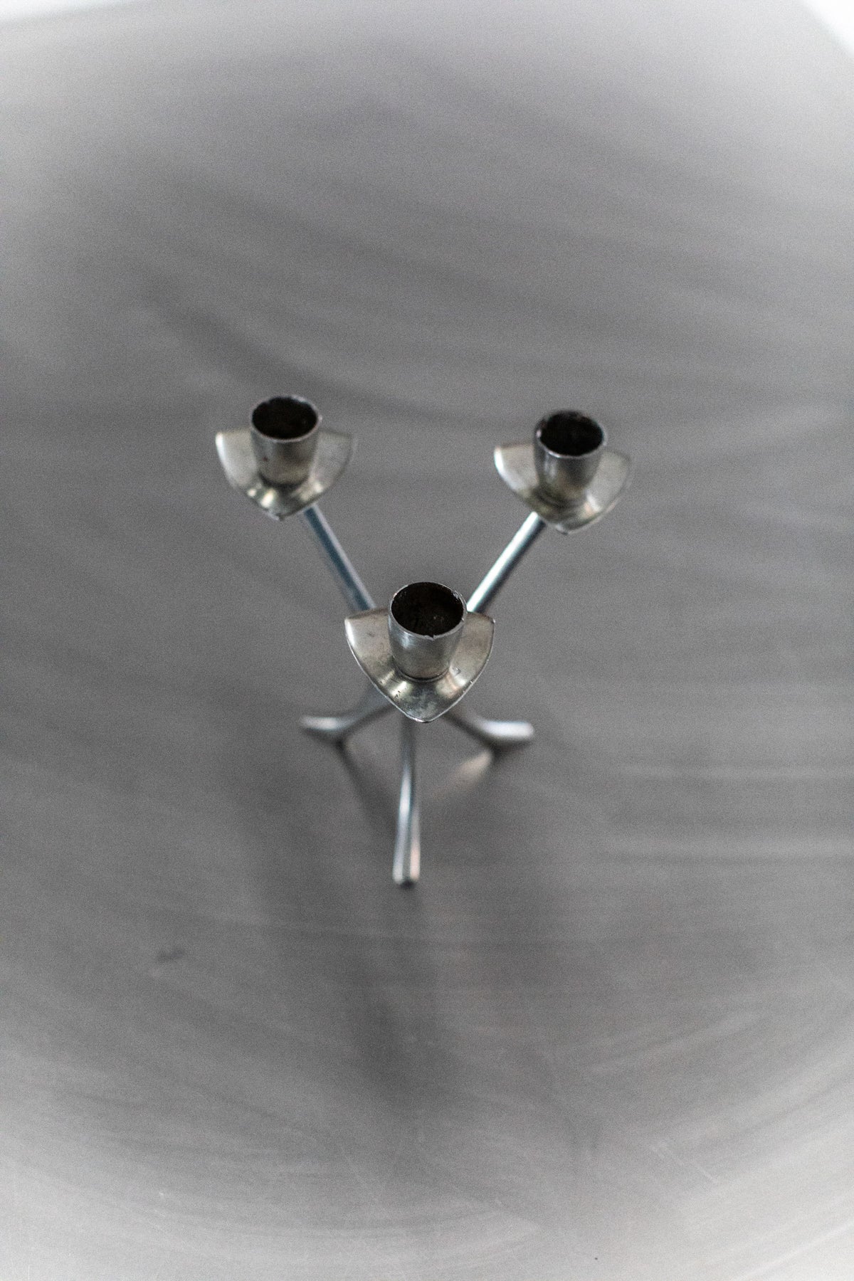 The Out For Lunch Steel Candleholder, featuring three vertically intersecting metal rods with cup-like tops, creates a modern vibe on a reflective surface. Its triangular design against gray gradients enhances its contemporary decor appeal.
