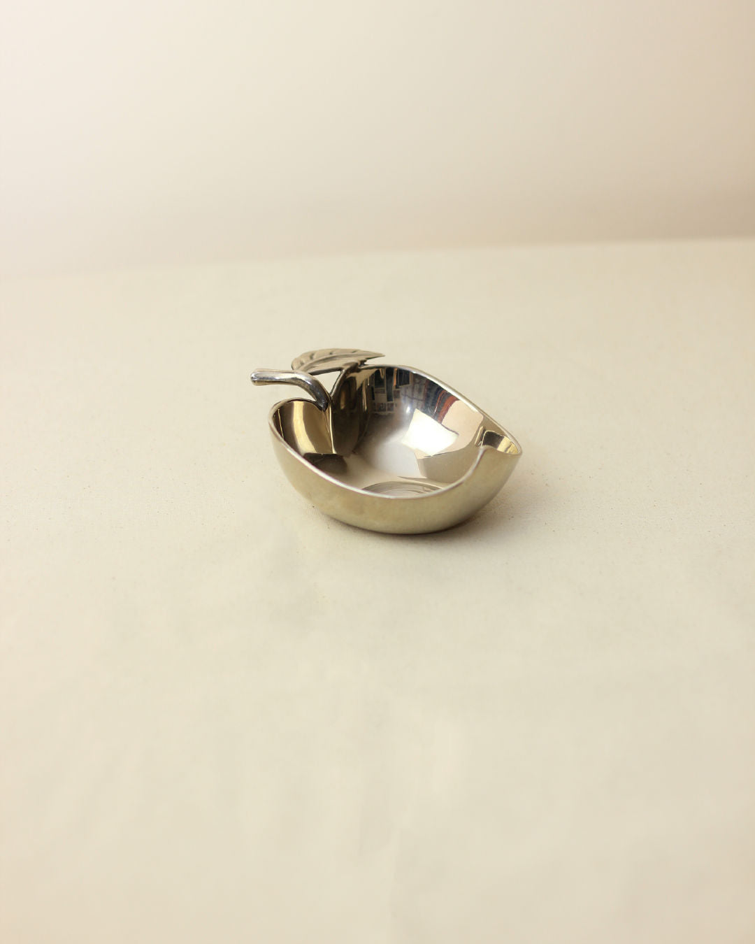 A shiny, small Decorative Apple Shaped Dish by Boga Avante Shop with a matching spoon rests on a plain, light-colored surface against a neutral background.