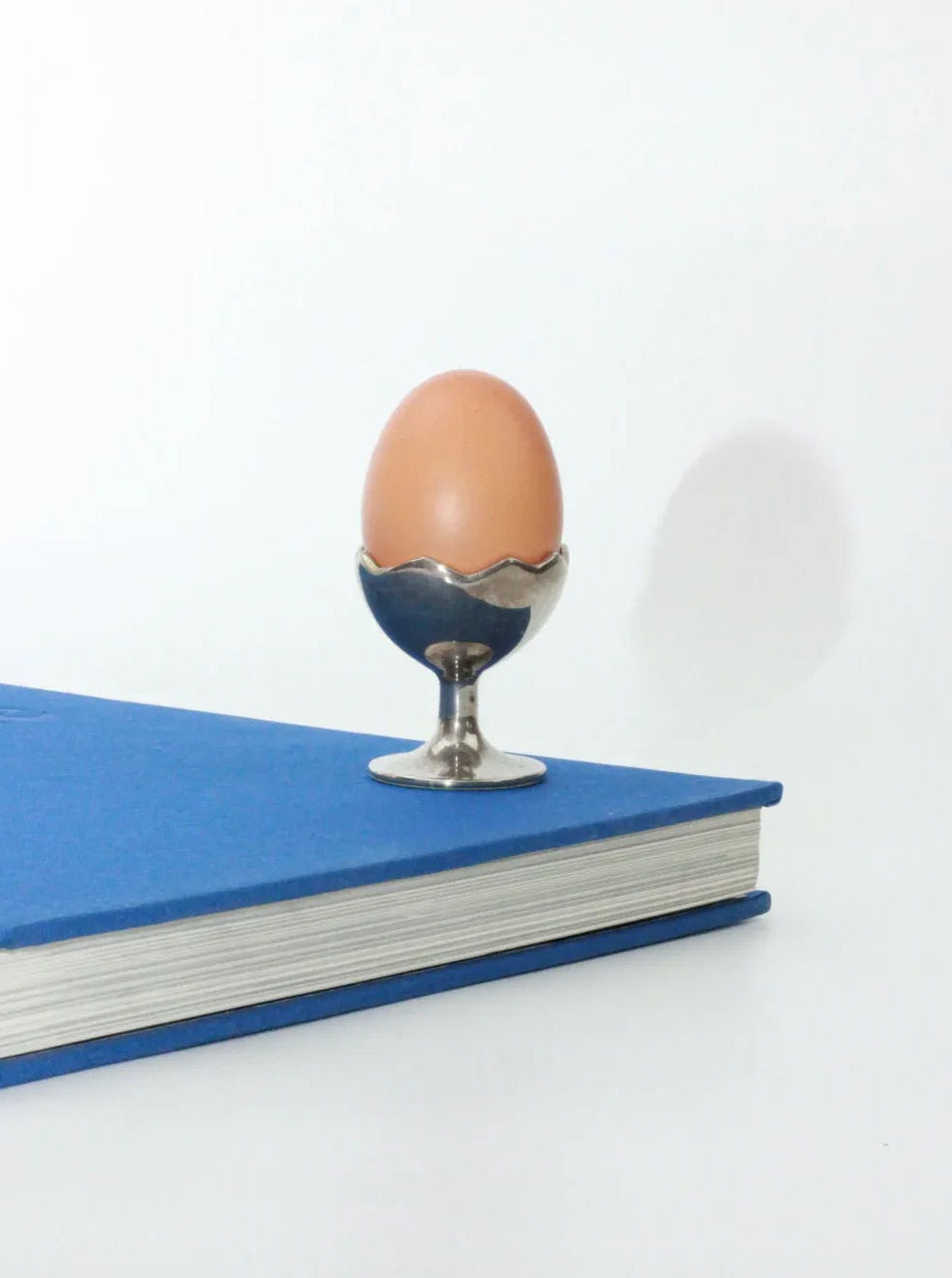 An Egg Cup from Boga Avante Shop sits upright in a silver alpaca egg cup placed on a closed blue book with a white background. The egg is centered on the book, and the minimalistic scene emphasizes simplicity.