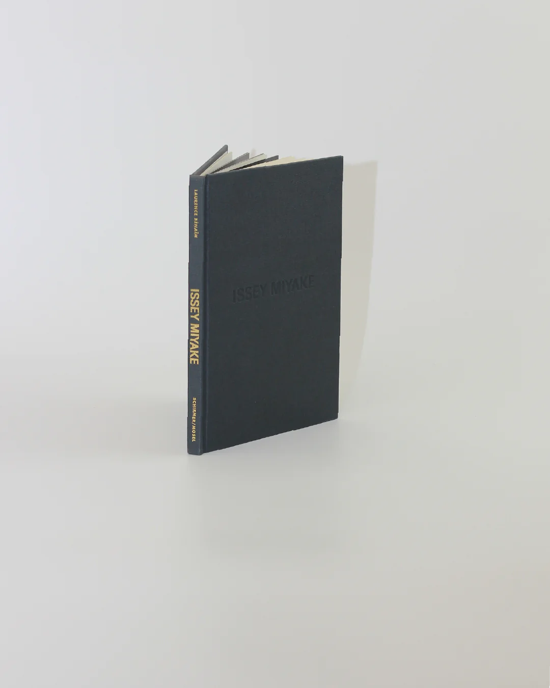 A hardcover book from Boga Avante Shop, titled "Issey Miyake," stands upright against a light gray background. Its black cover features the name "Issey Miyake" embossed in yellow on the spine. The book is slightly open, offering a glimpse into Miyake's exploration of contemporary fashion and innovative pleating techniques.