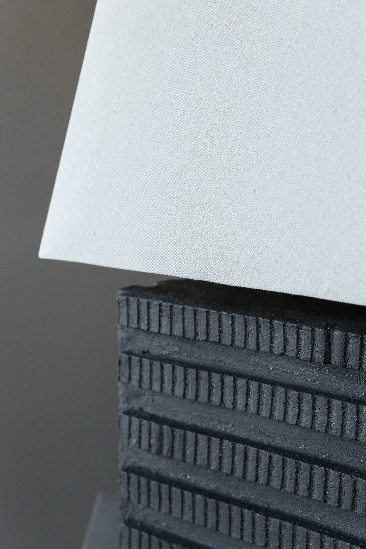 Close-up of the Akune Ceramic Lamp - Number One by Studio OSKLO, featuring a textured black rectangular base with horizontal ridges and a light gray fabric lampshade, inspired by post-Bauhaus brutalism.