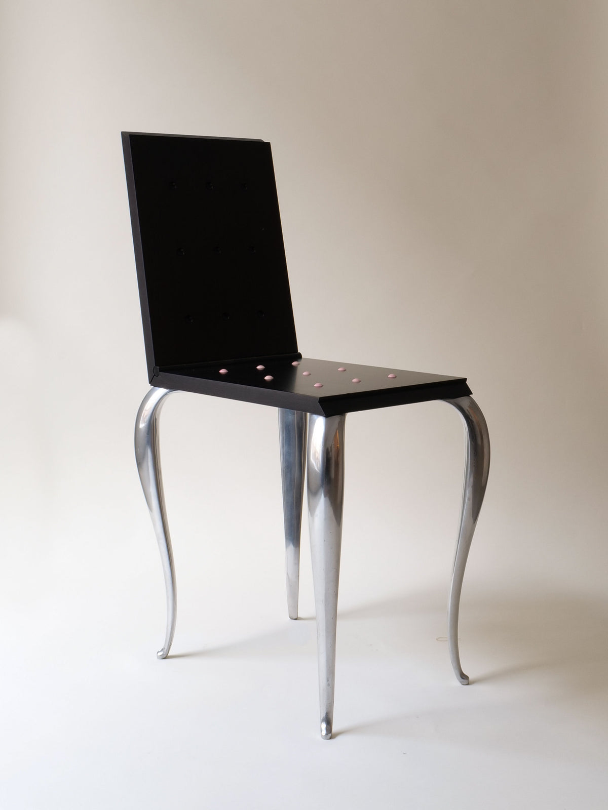 The "Lola Mundo Chair" by Philippe Starck for Collection apart (1984) boasts a black square seat and backrest adorned with small pink dots. Its sleek, silver metallic legs curve outward, exuding a Starck-inspired design, making it an artistic and collectible piece against a plain white backdrop.