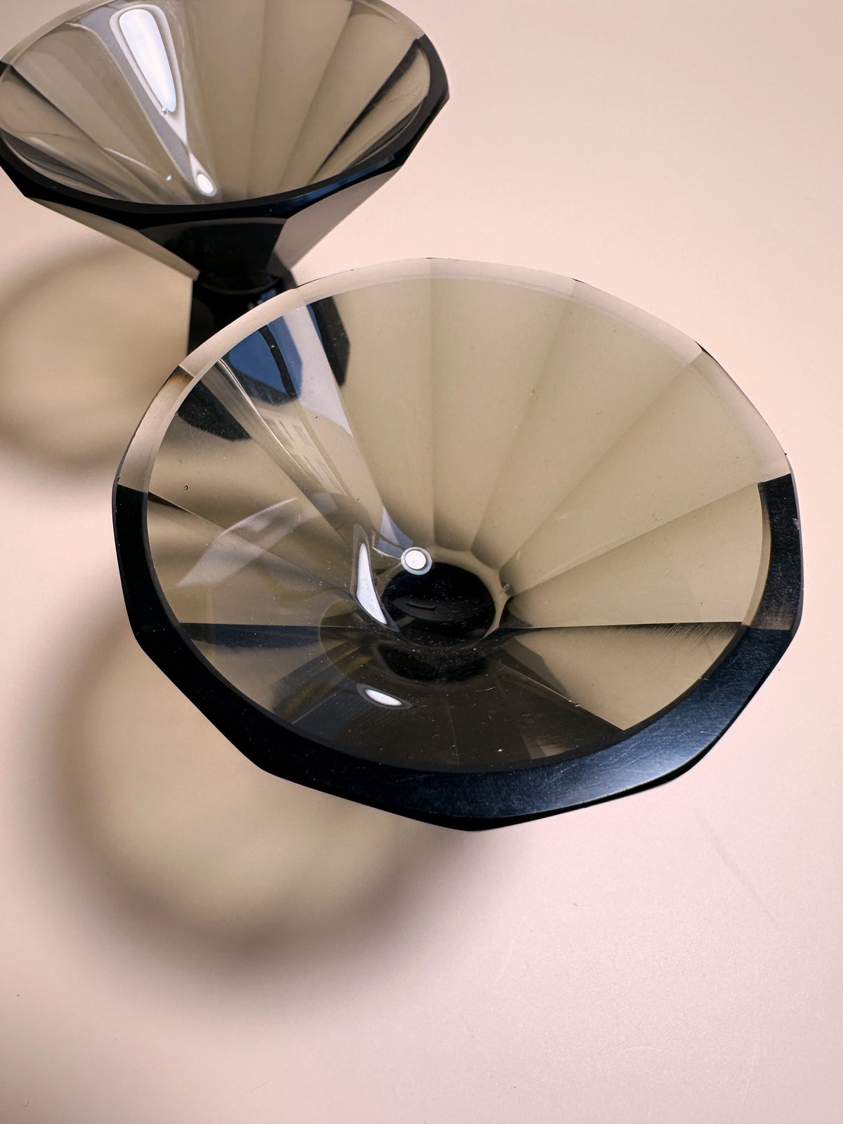 A close-up view of two translucent, dark gray, geometric-shaped pieces named "1930s Art Deco Facetted Liqueur Glasses" by Dodo Vintage. Crafted from smoked glass and resting on a light beige surface, one glass is slightly behind and to the side of the other, casting a shadow that enhances their handmade elegance.