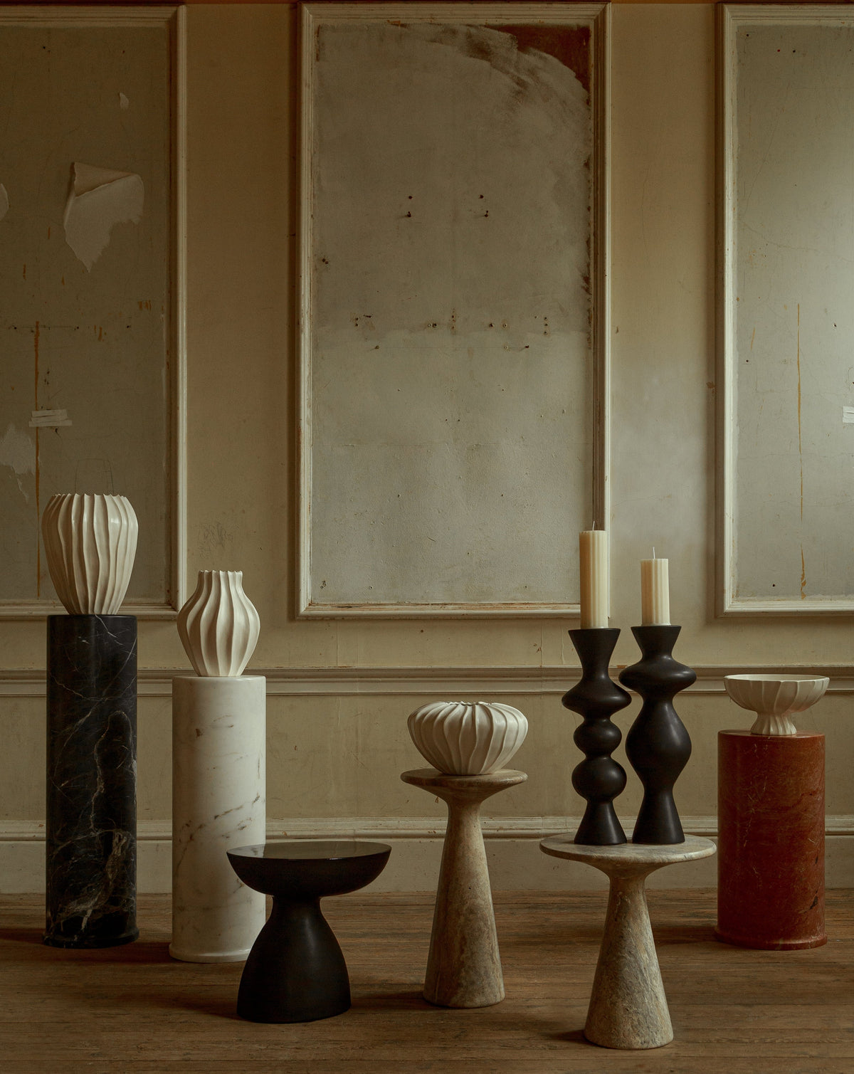 A rustic room featuring a wooden floor and neutral-toned walls displays a ceramic collection, including artistic sculptures and candle holders on pedestals of differing heights. The Medium Cloak Vase by Ransom & Dunn, handmade in Portugal, complements the decor with its abstract shapes in black, white, and earthy tones.