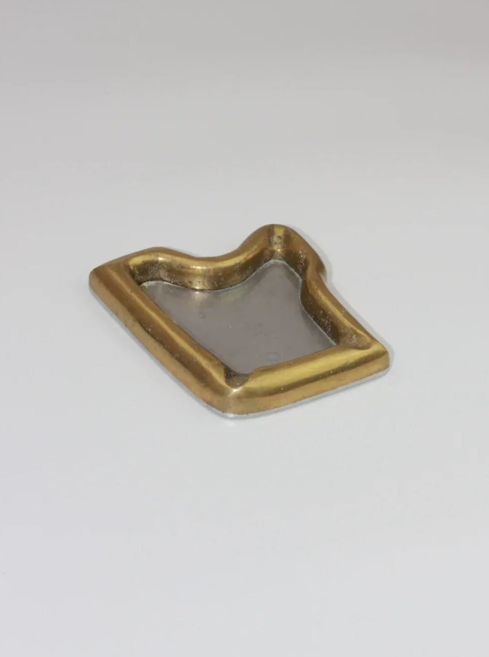 A rectangular, gold-framed Brutalist Ashtray by Boga Avante Shop with raised edges lies flat on a neutral surface. The center of the ashtray is a silver-colored metal, contrasting with the gold border. The slightly uneven edges give this vintage item a handcrafted appearance reminiscent of Brutalist style.