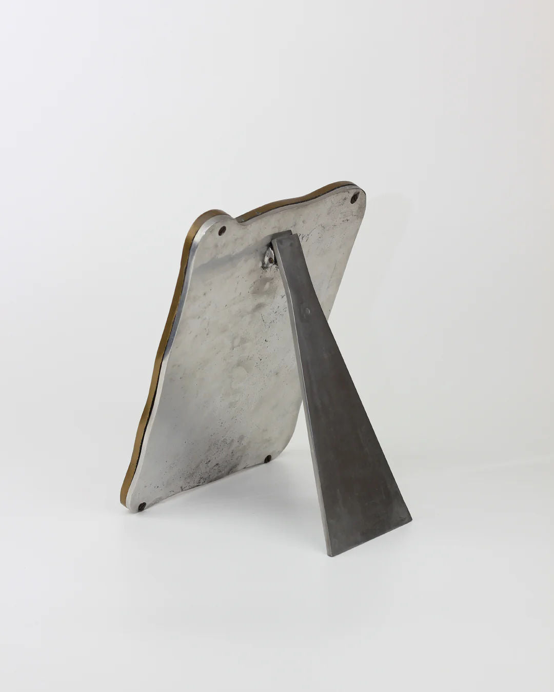 The Brutalist Mirror from Boga Avante Shop features a metal triangular base connected to a larger irregularly shaped flat piece with an industrial finish, smooth texture, slight patina, and two small holes at the top, all set against a white background.