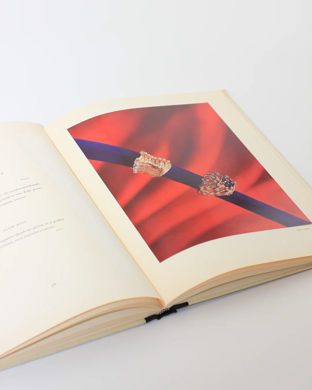 An open book titled "Dalí: A Study of his Art-in-Jewels" by Boga Avante Shop, lies on a light surface, showing two gold rings against a red and blue backdrop. The nearby text page partially visible hints at the artistic legacy it explores.