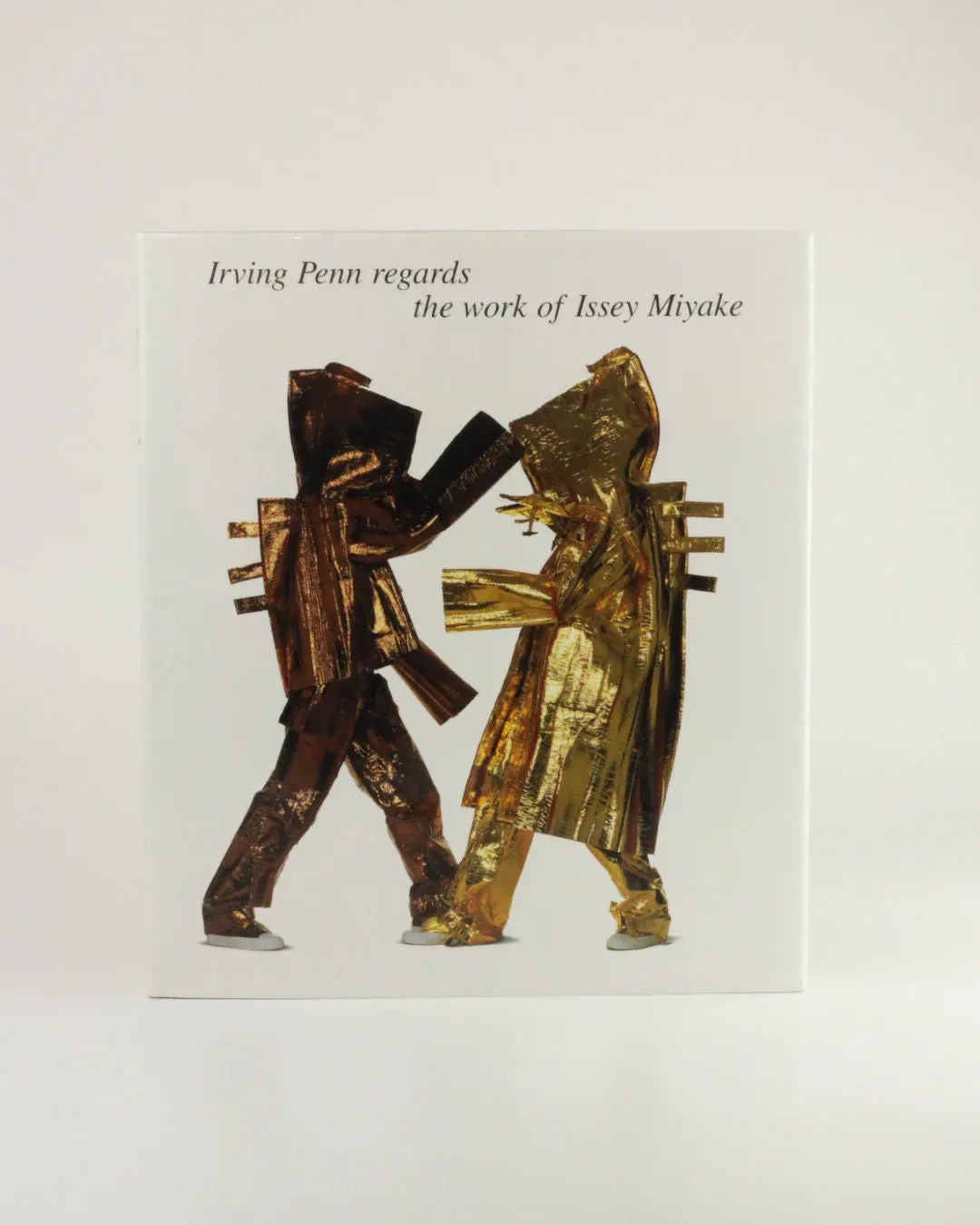 Photograph of two models dressed in metallic, sculptural outfits posed as if interacting. The text at the top reads "Irving Penn regards the work of Issey Miyake." The plain white background highlights the striking, artistic fashion designs captured in this exceptional fashion photography by Boga Avante Shop.