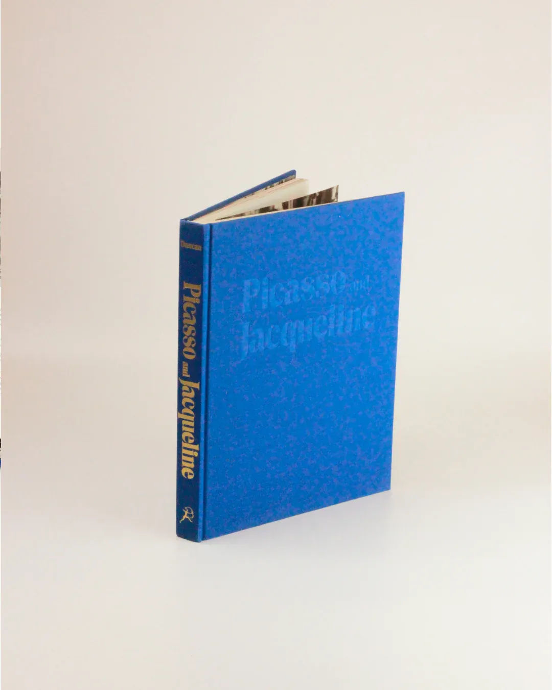 A blue hardcover book titled "Picasso and Jacqueline" from Boga Avante Shop stands upright against a plain white background. The title, printed in gold and white text along the spine, is faintly visible on the front cover. The book is slightly open, offering an intimate portrait through its revealed pages.