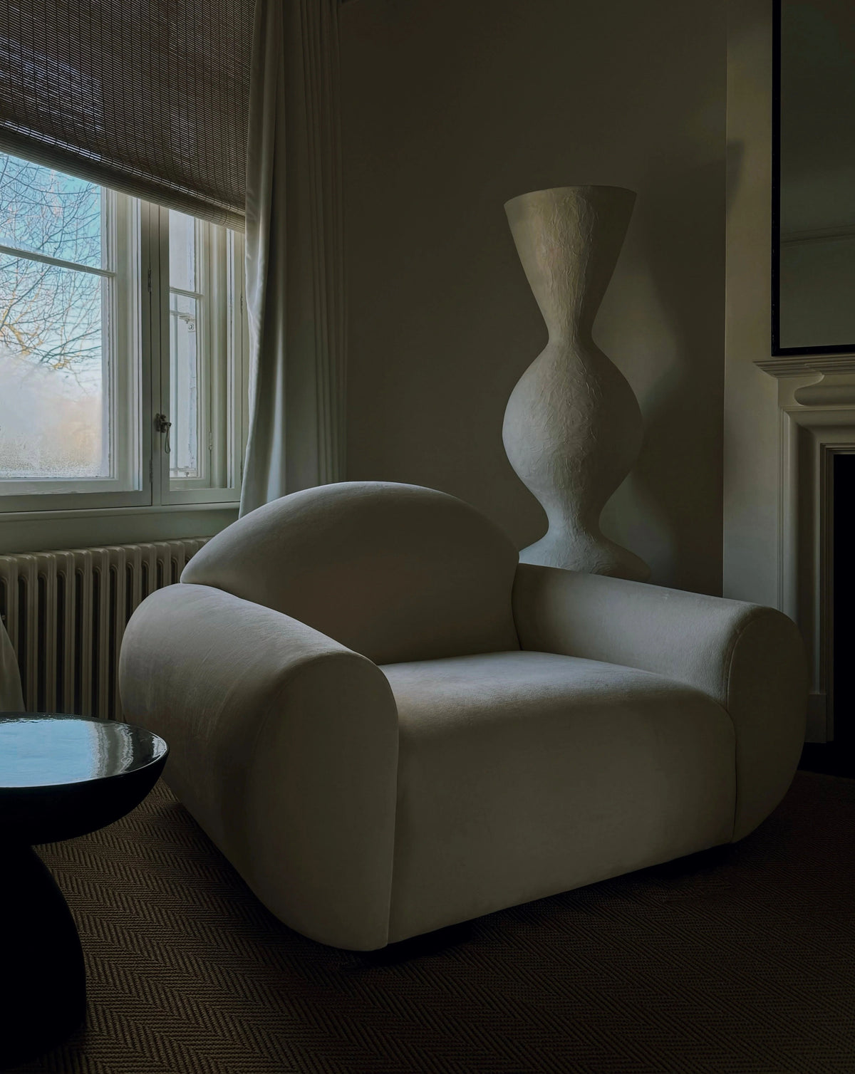 A cozy living room showcases the "Sparta" Armchair by Ransom & Dunn, featuring a large and soft design upholstered in luxurious alpaca wool. Beside it sits a tall, sculptural vase paired with a small table. A window dressed with sheer curtains lets natural light filter through, casting a gentle glow over the neutral-toned space.