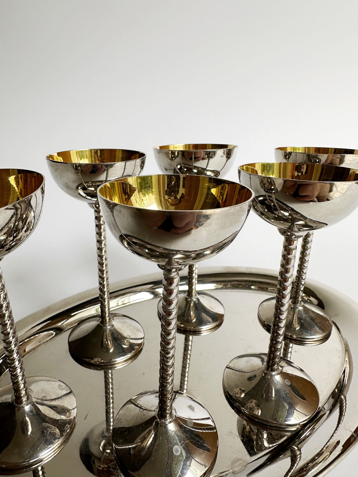 Introducing the **Art Nouveau liqueur goblet set** by **Dodo Vintage**: a collection of six silver-plated, gold-lined goblets that evoke the elegance of Art Nouveau design with their intricate, twisted stems. Each goblet is perfectly arranged on a matching silver tray. The shiny surfaces and exquisite craftsmanship not only reflect light beautifully but also add a touch of sophistication to any setting.