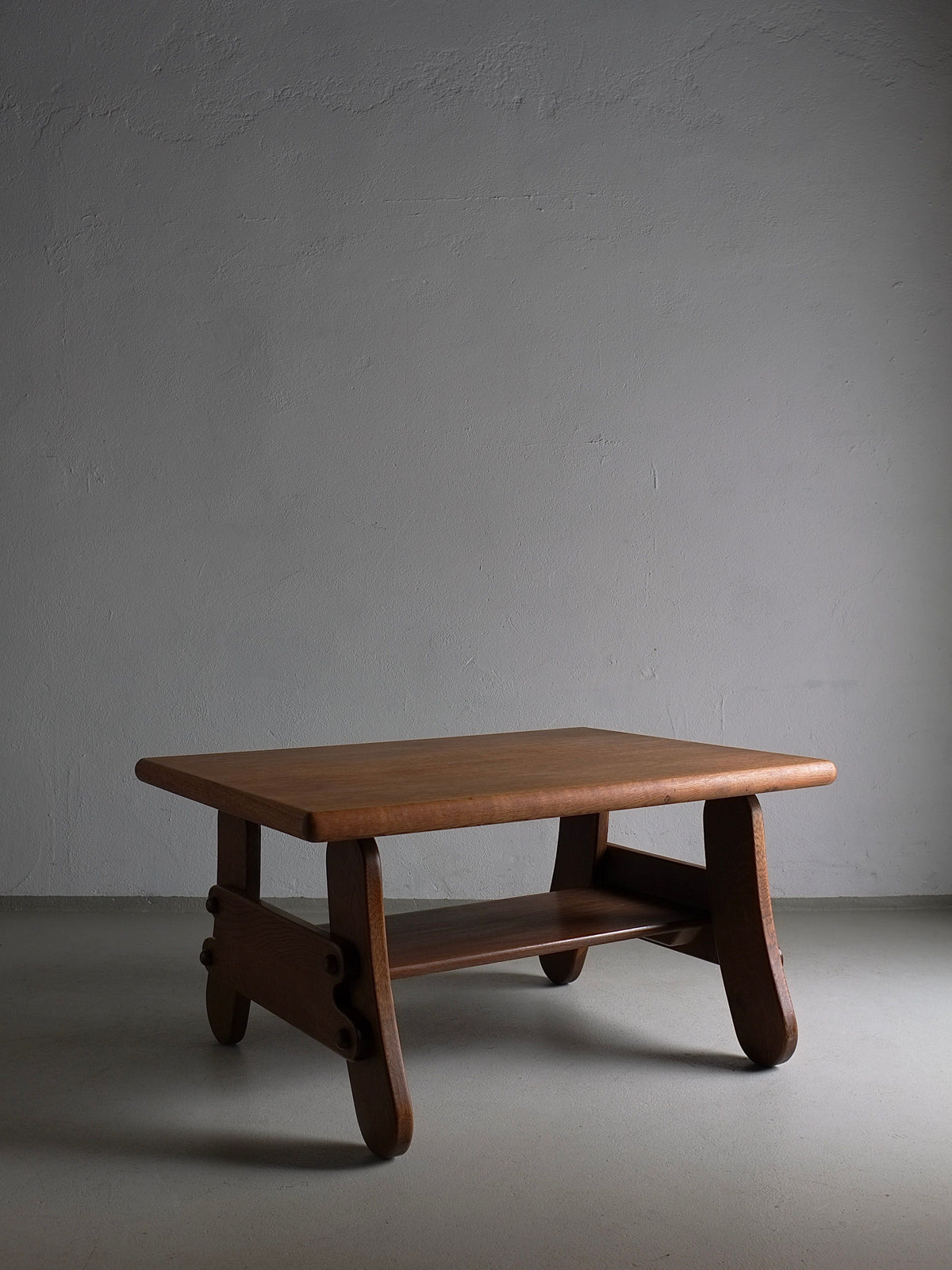 A vintage coffee table with a rectangular top and curved legs stands against a plain gray backdrop. Crafted in a 1970s design, the Brutalist Oak Coffee Table | Netherlands | 1970s by Veter Vintage has a small lower shelf for extra storage. The lighting casts subtle shadows, highlighting the table's smooth finish and simple yet elegant design.