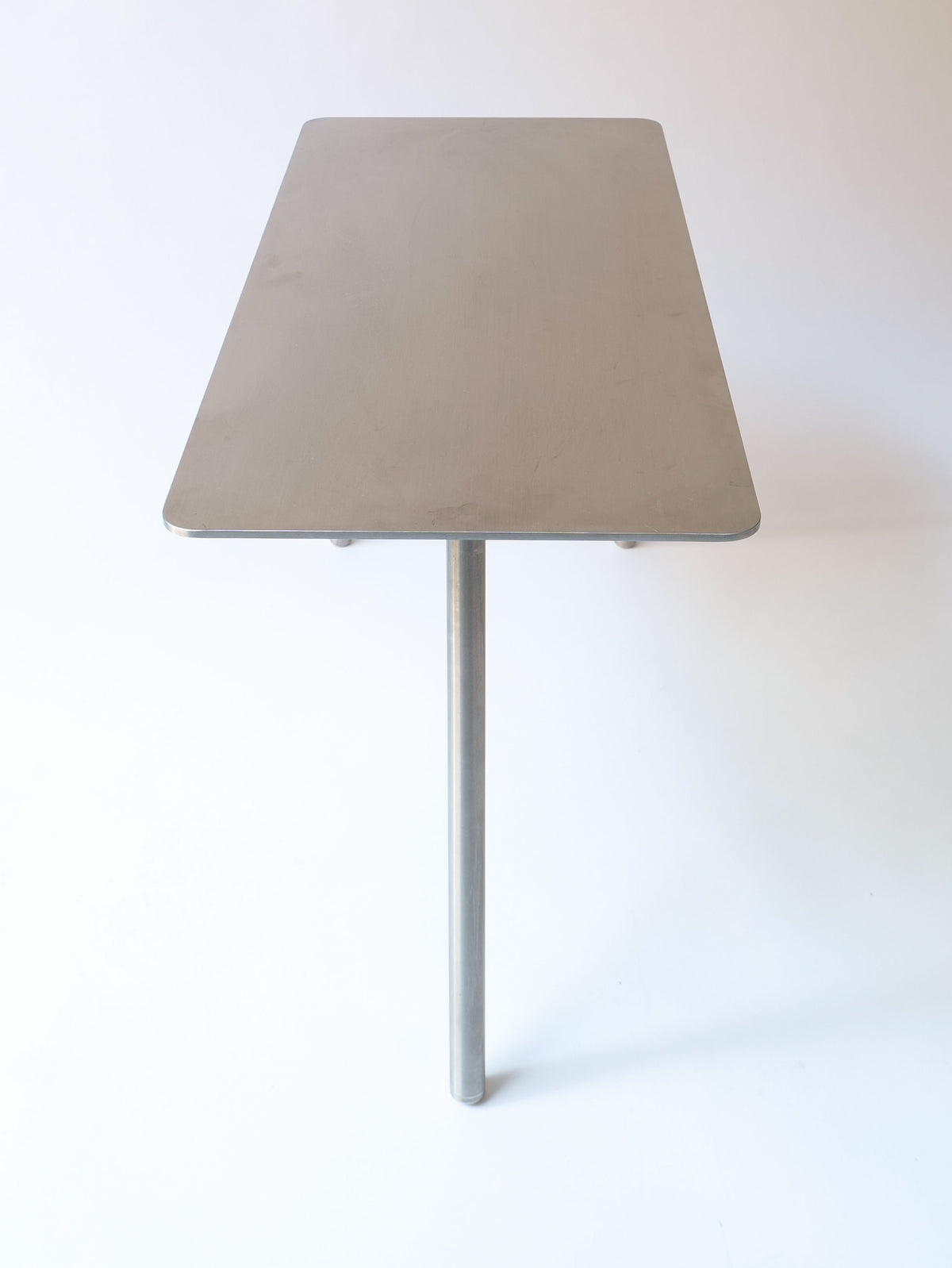 A minimalist rectangular Stick Table Jesper Morrison 2000 with a single central cylindrical leg, positioned against a plain white background. Designed by Jasper Morrison for Collection apart, the tabletop has a smooth, flat surface with slightly rounded edges and is crafted from elegant Italian stainless steel.