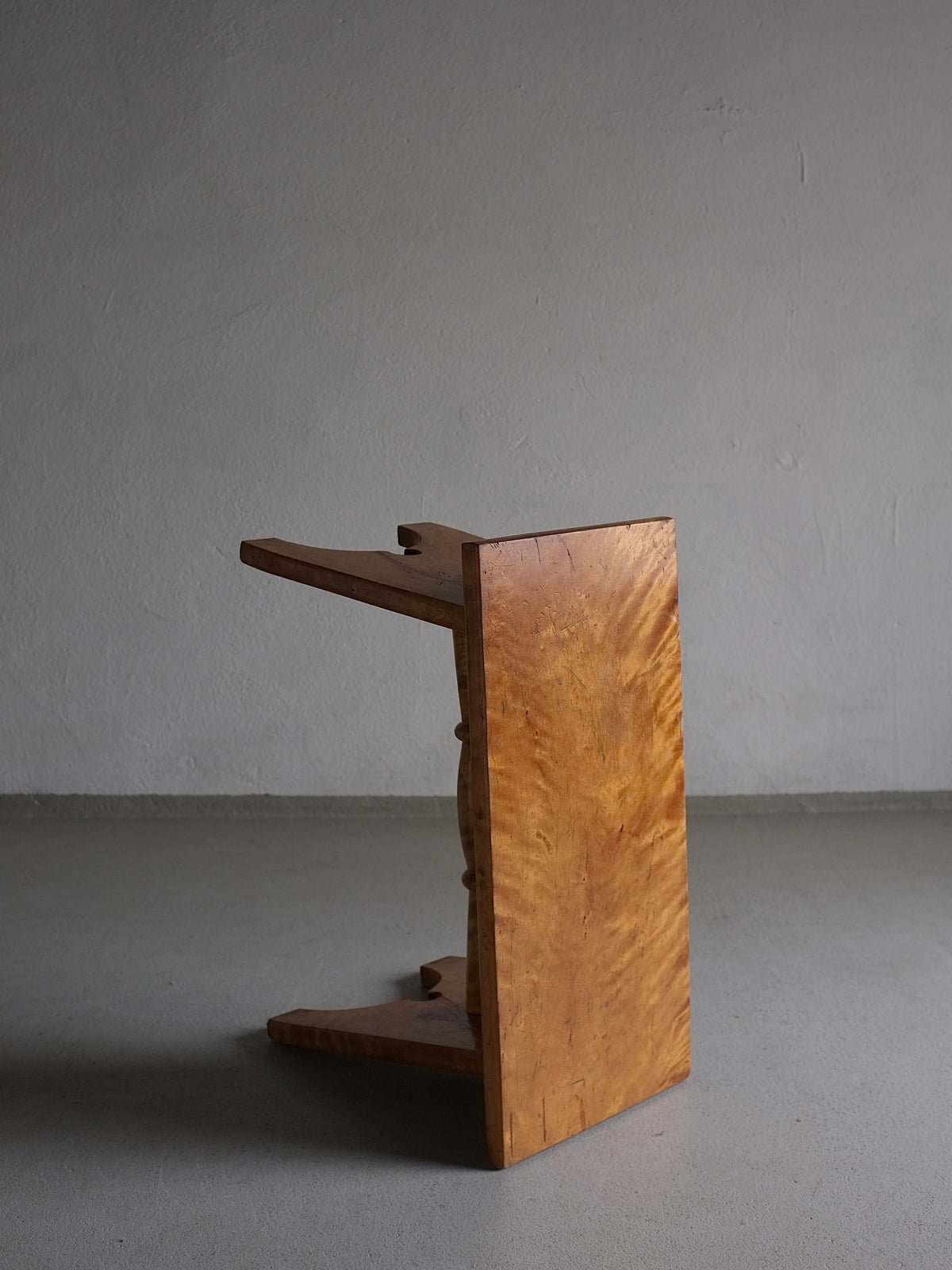 A Rustic Solid Karelian Birch Stool | Latvia | 1950s by Veter Vintage, crafted from solid Karelian birch wood, is positioned sideways, resting on its side against a bare, light gray wall. The stool has a simple design with a smooth finish, showcasing the wood grain texture. The setting is minimalistic, with no additional objects or decorations in the scene.