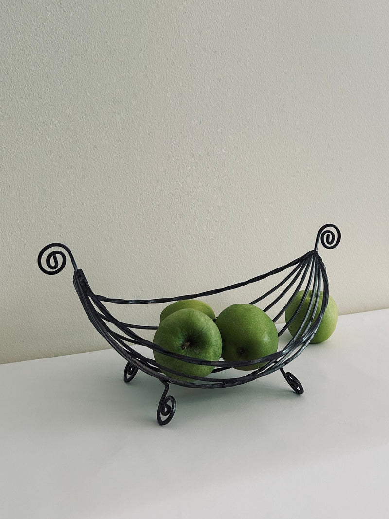 The Swirl Fruit Basket by Flock Studio, crafted from metal wire, cradles green apples on a white surface against a plain white wall. With its elegant curved design and swirled ends, it not only delights the eye but also ensures excellent air circulation.