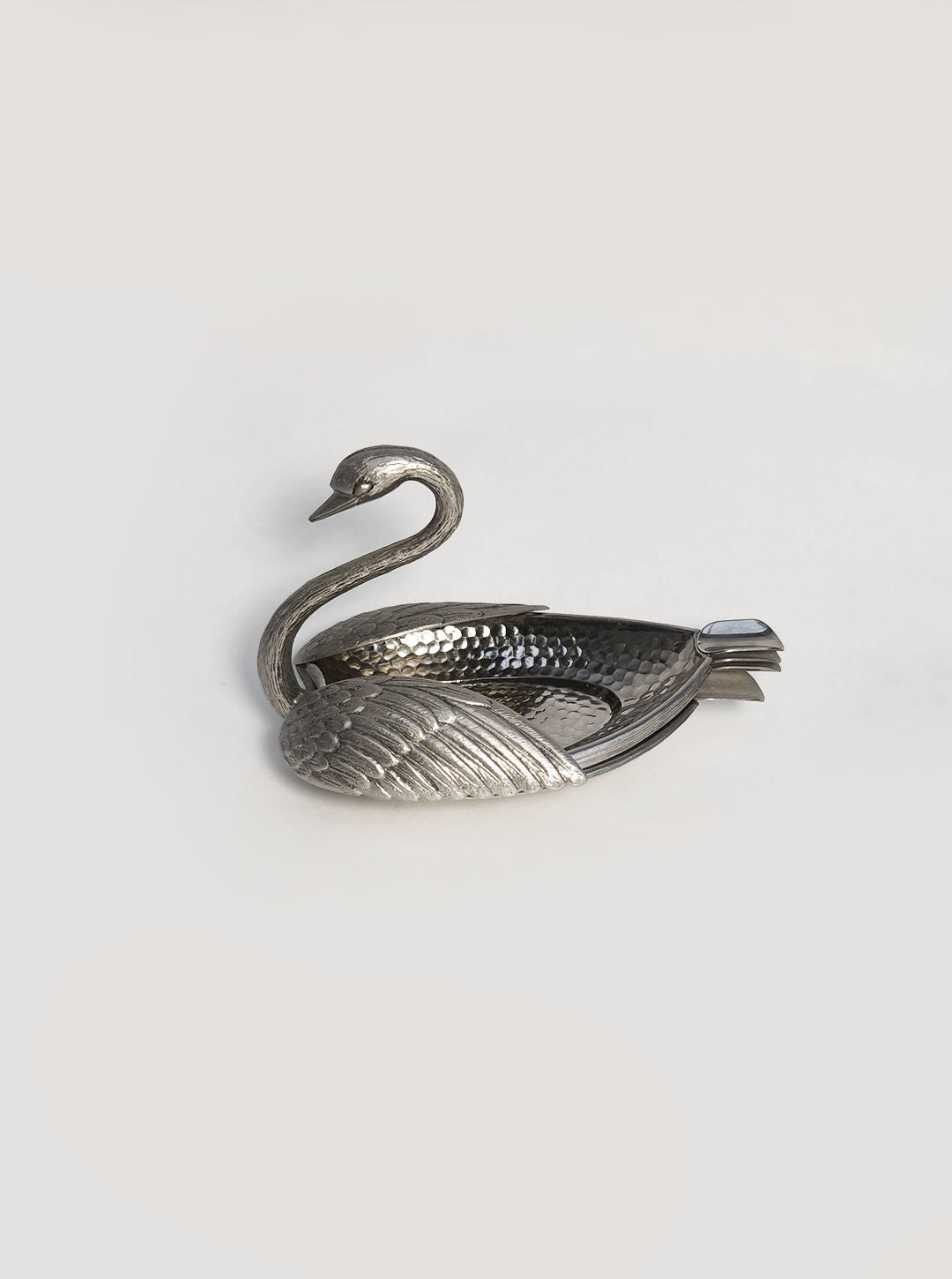 The Les Objoies Swan Ashtray is depicted with a textured, silver swan-shaped design. Its gracefully bent head and wings create compartments reminiscent of an elegant metal napkin holder against a plain white backdrop.