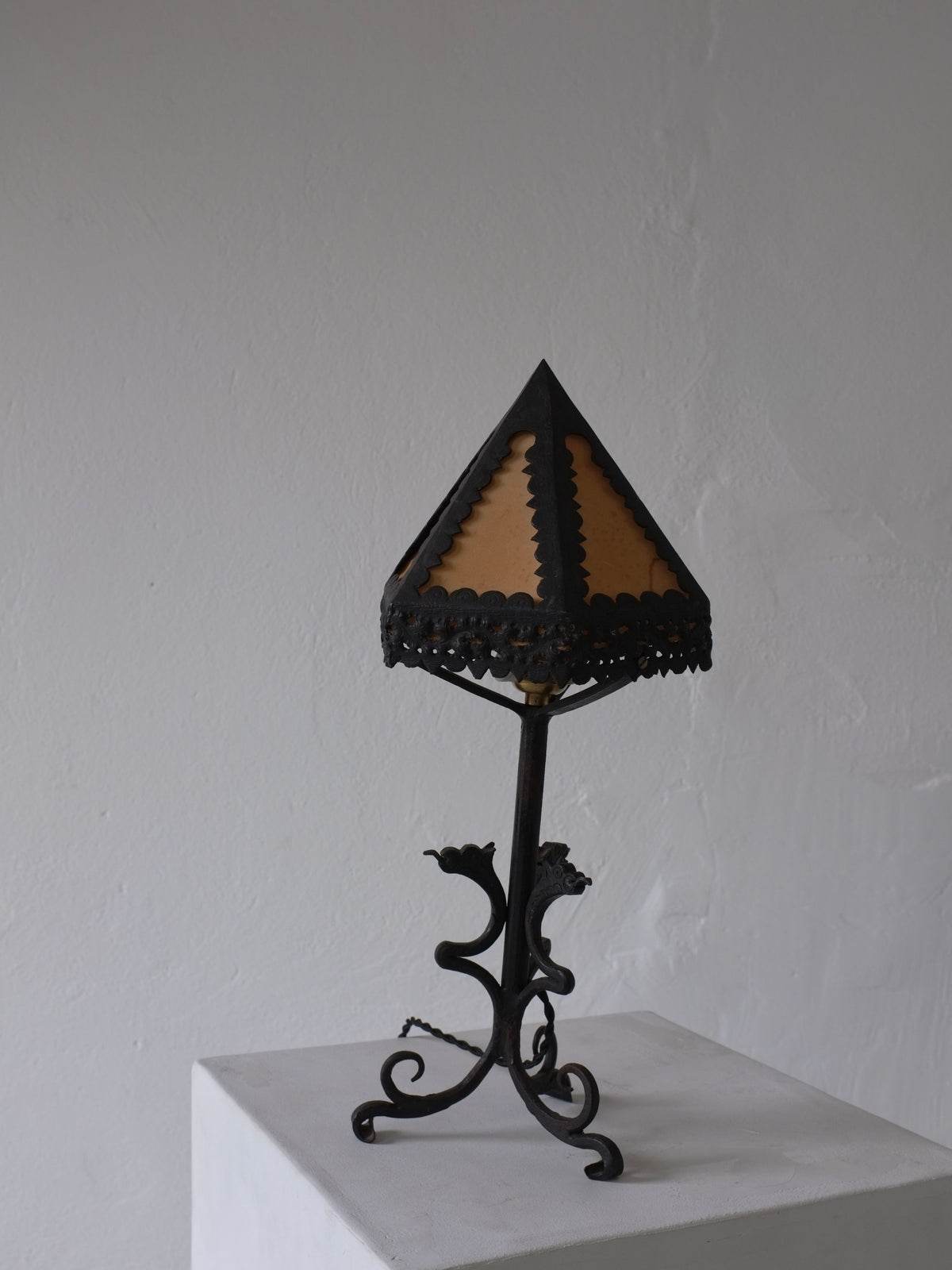 The Veter Vintage Wrought Iron Dragon Table Lamp, Italy 1910s, features ornate dragon details, a black base, and a triangular tan silk shade, displayed on a white pedestal against a light gray wall.